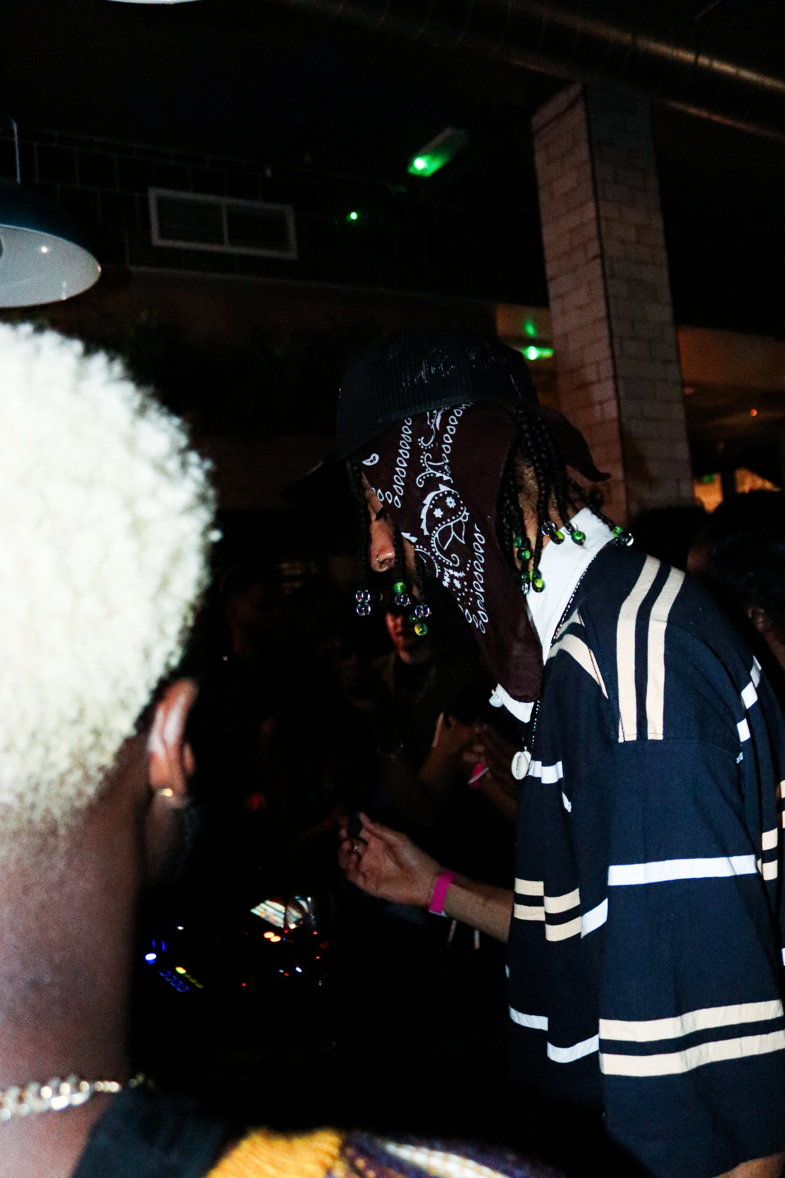A person with braided hair and a bandana on their head, wearing a striped shirt and colorful beads, appears to be in a dimly lit nightclub or party setting with a DJ mixer in the background.
