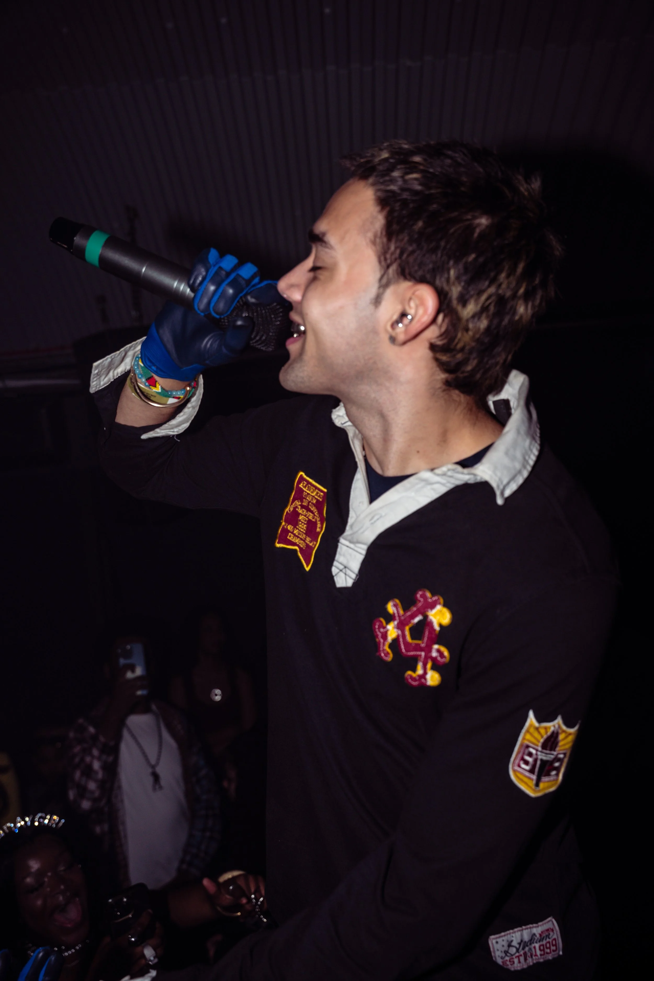 A young man with dark hair singing into a microphone at a crowded event, wearing a black jacket with patches and colorful bracelets.