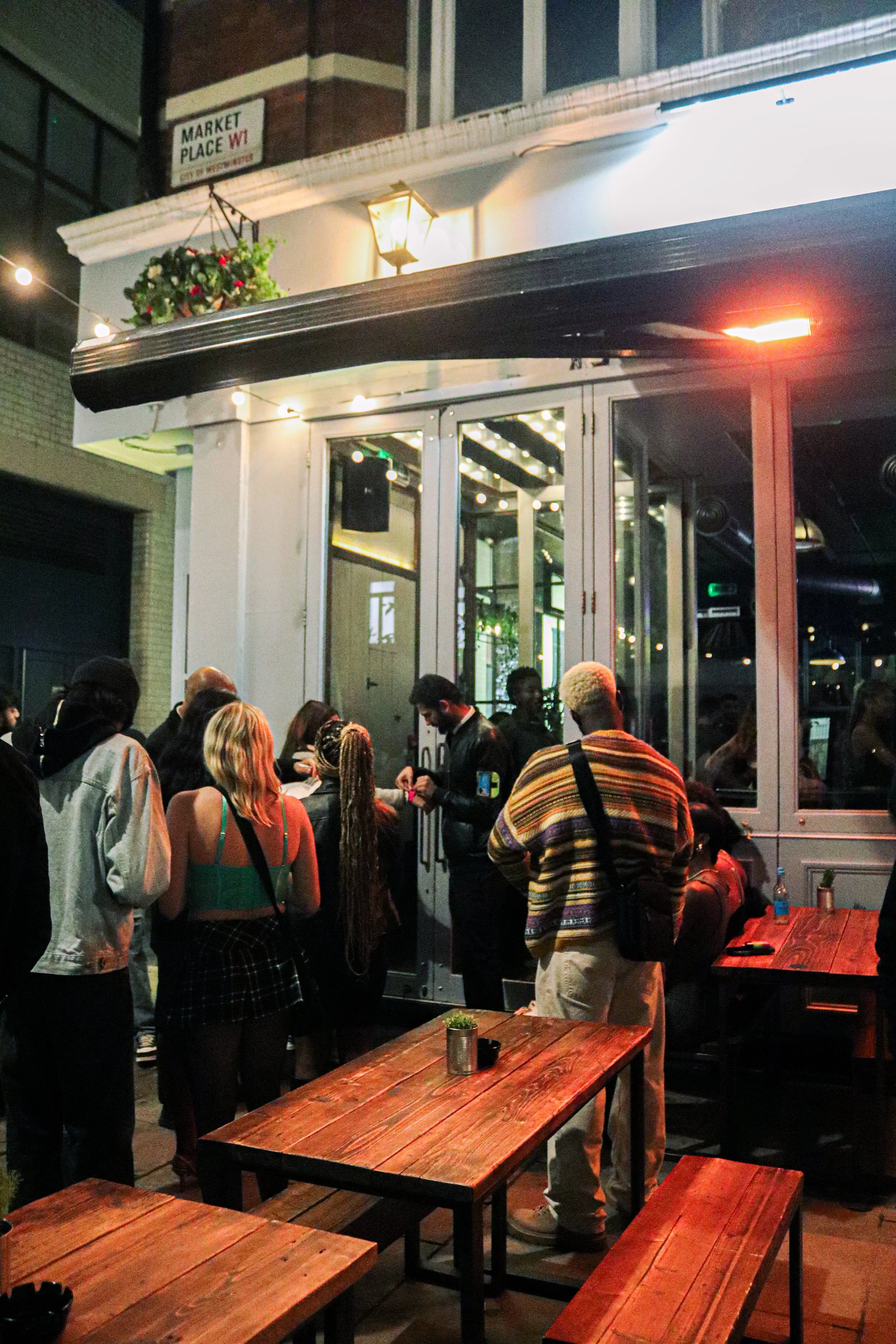 People in line inside a restaurant with wooden tables and a glass window. Nighttime, warm lighting, and some people are waiting or ordering food.