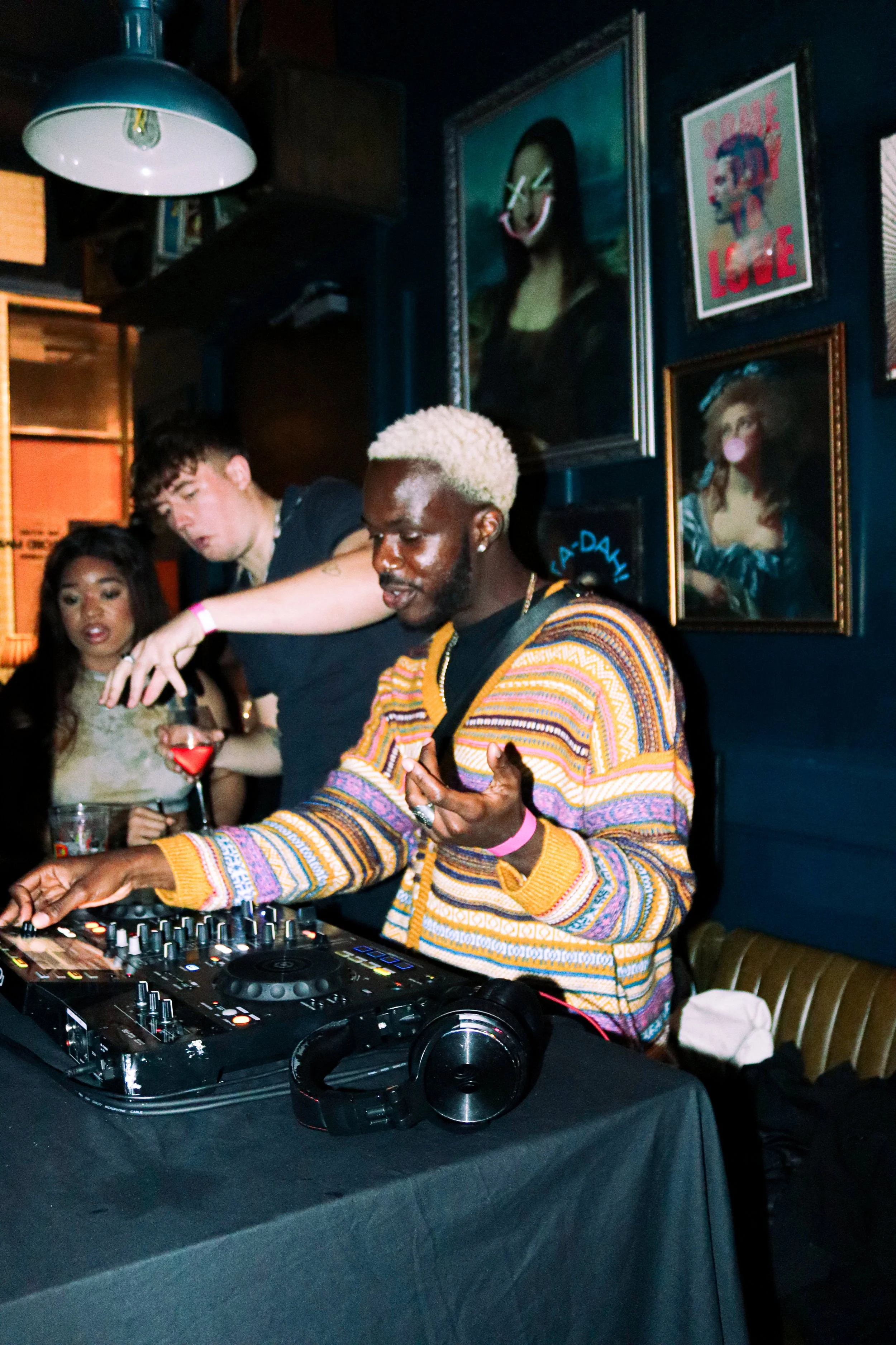 A DJ with platinum blonde hair and a multi-colored sweater performs in a dimly lit room. Three people are visible in the background, one holding a drink. The walls are decorated with various framed artworks, including a portrait of Mona Lisa with cro