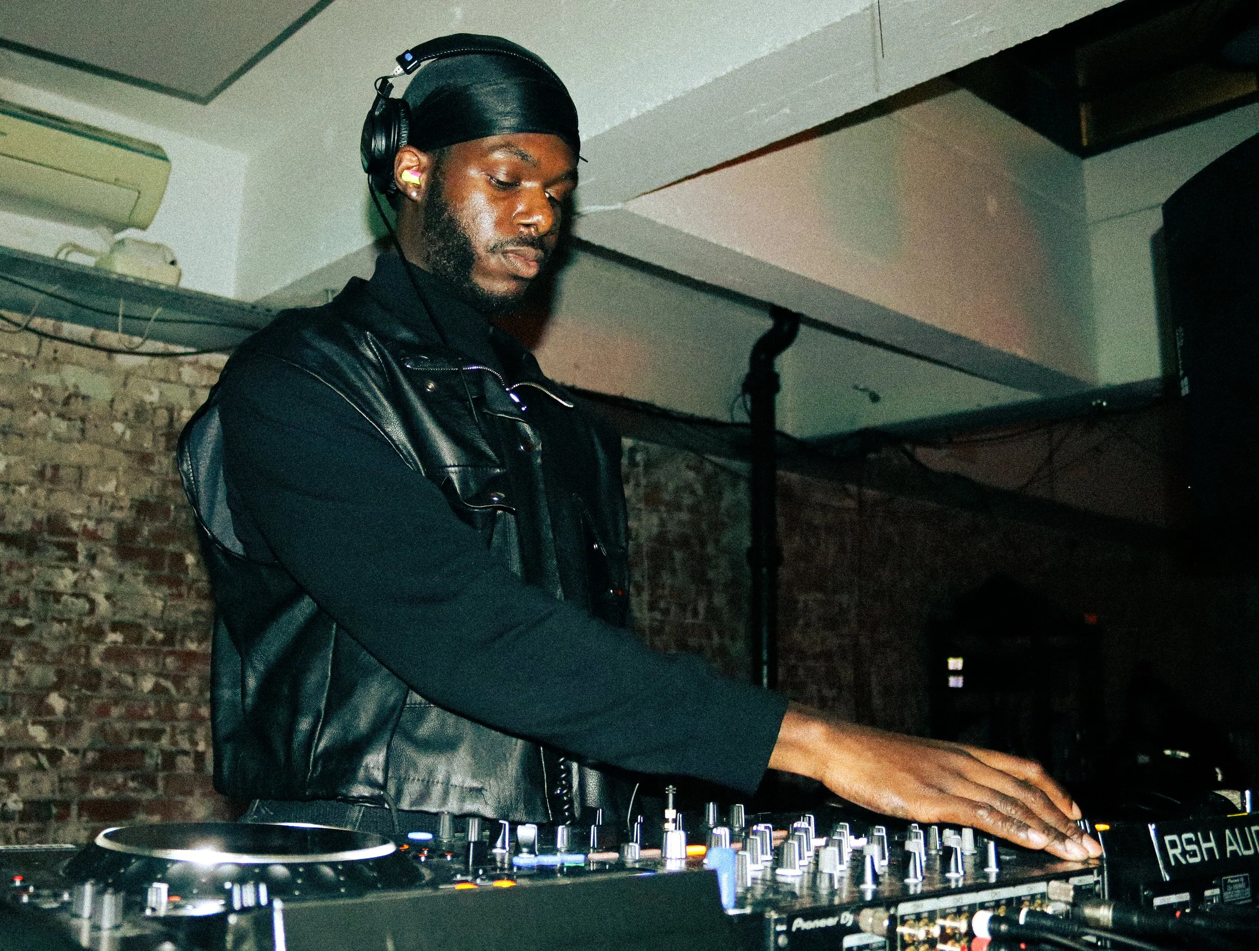 A DJ with dark skin wearing a black leather jacket, black head wrap, and yellow earplugs operates a DJ mixer and turntable in a dimly lit venue with brick walls.