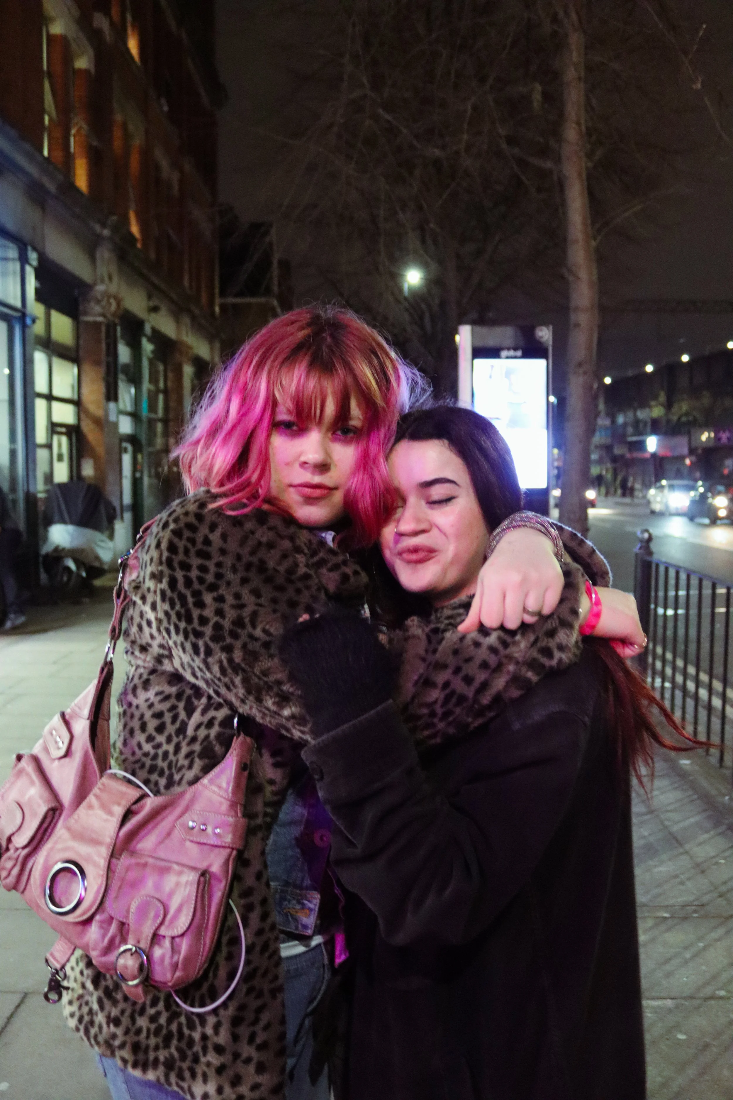 Two women hugging on a city sidewalk at night, one with pink hair and a pink bag, the other with dark hair and a dark coat, with streetlights and a building in the background.