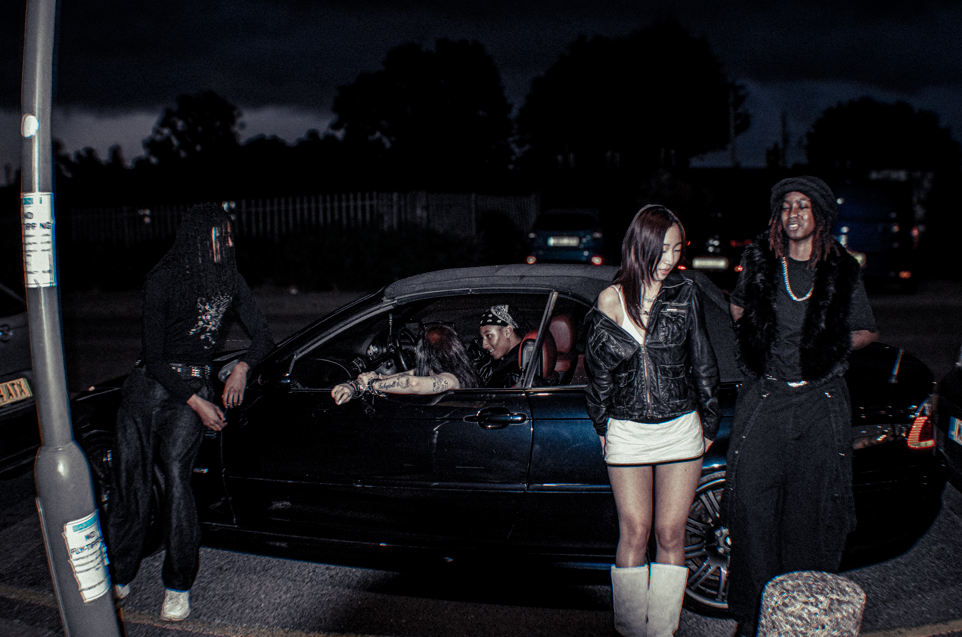 Group of young people near a black convertible car at night, with three standing outside and two inside the car, all dressed casually.