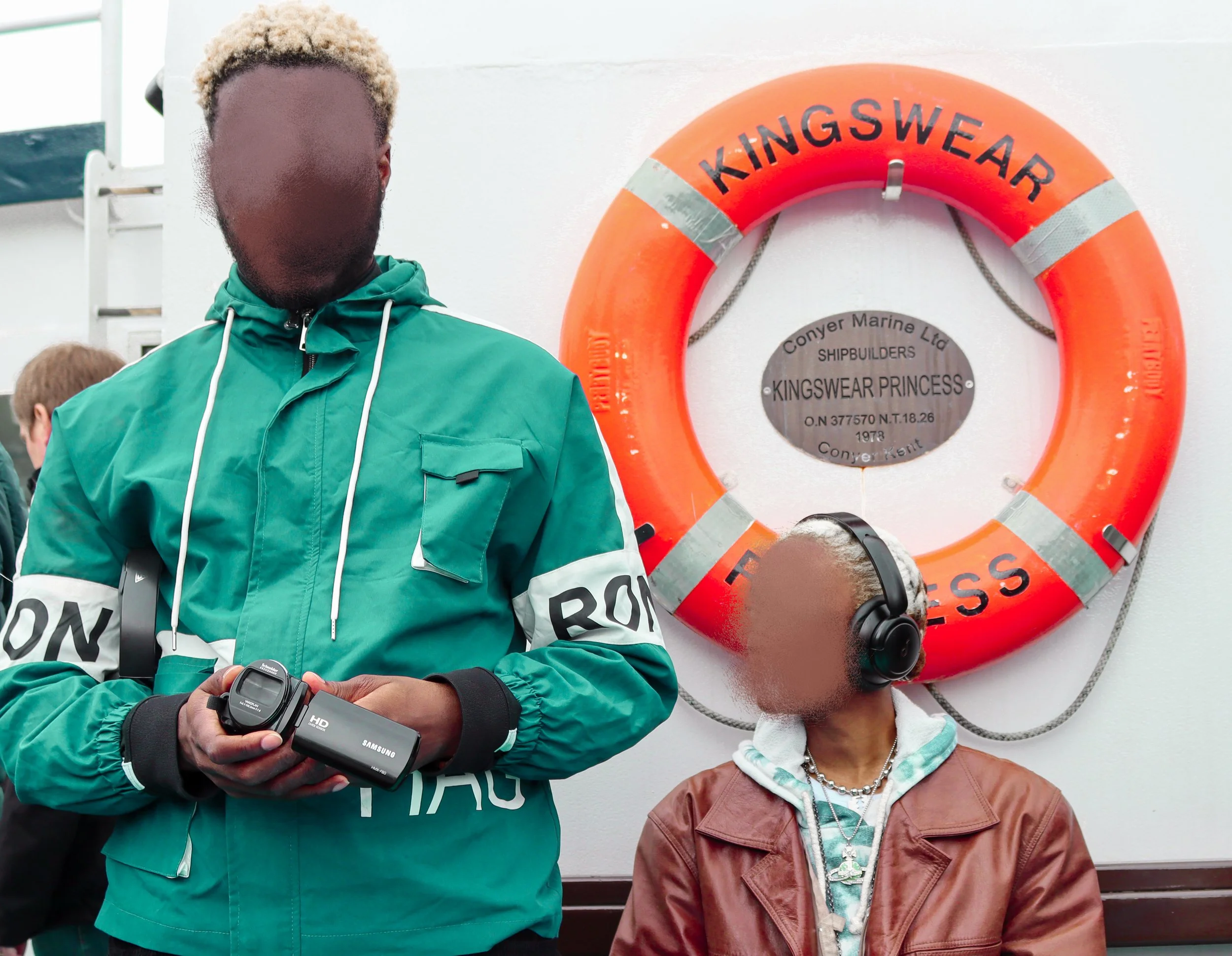 A person with curly blonde hair wearing a green jacket, holding a Samsung camera, standing next to a person in a brown leather jacket and a necklace with an anchor pendant, with a bright orange lifebuoy labeled 'KINGSWEAR' and a plaque in the backgro