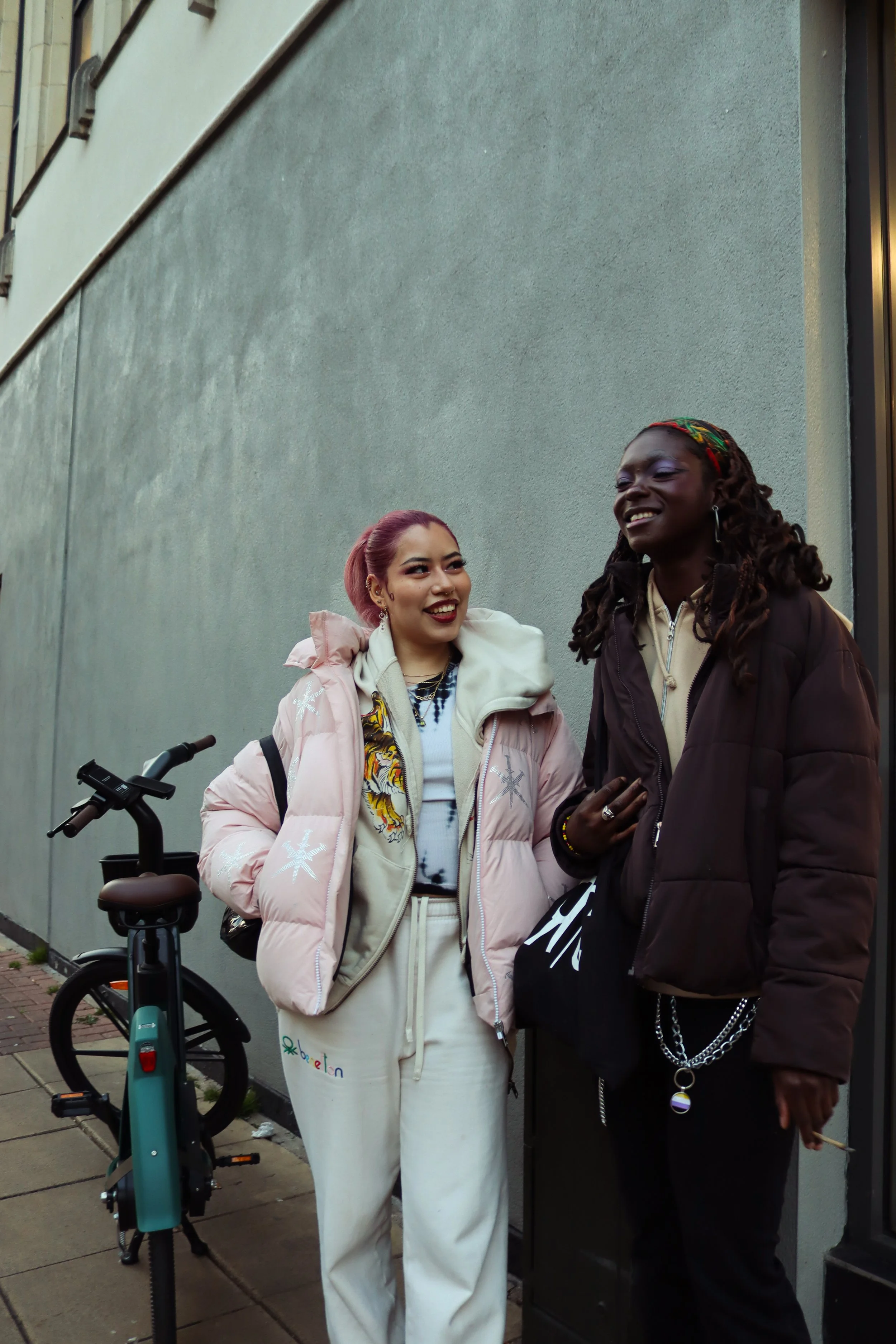 Two women are standing and smiling on a sidewalk against a plain gray wall. One woman has purple hair, a light pink puffer jacket, and white sweatpants with the word Google on them. The other woman has dark skin, dreadlocks, gold hoop earrings, a dark brown jacket, and a facial expression of joy.