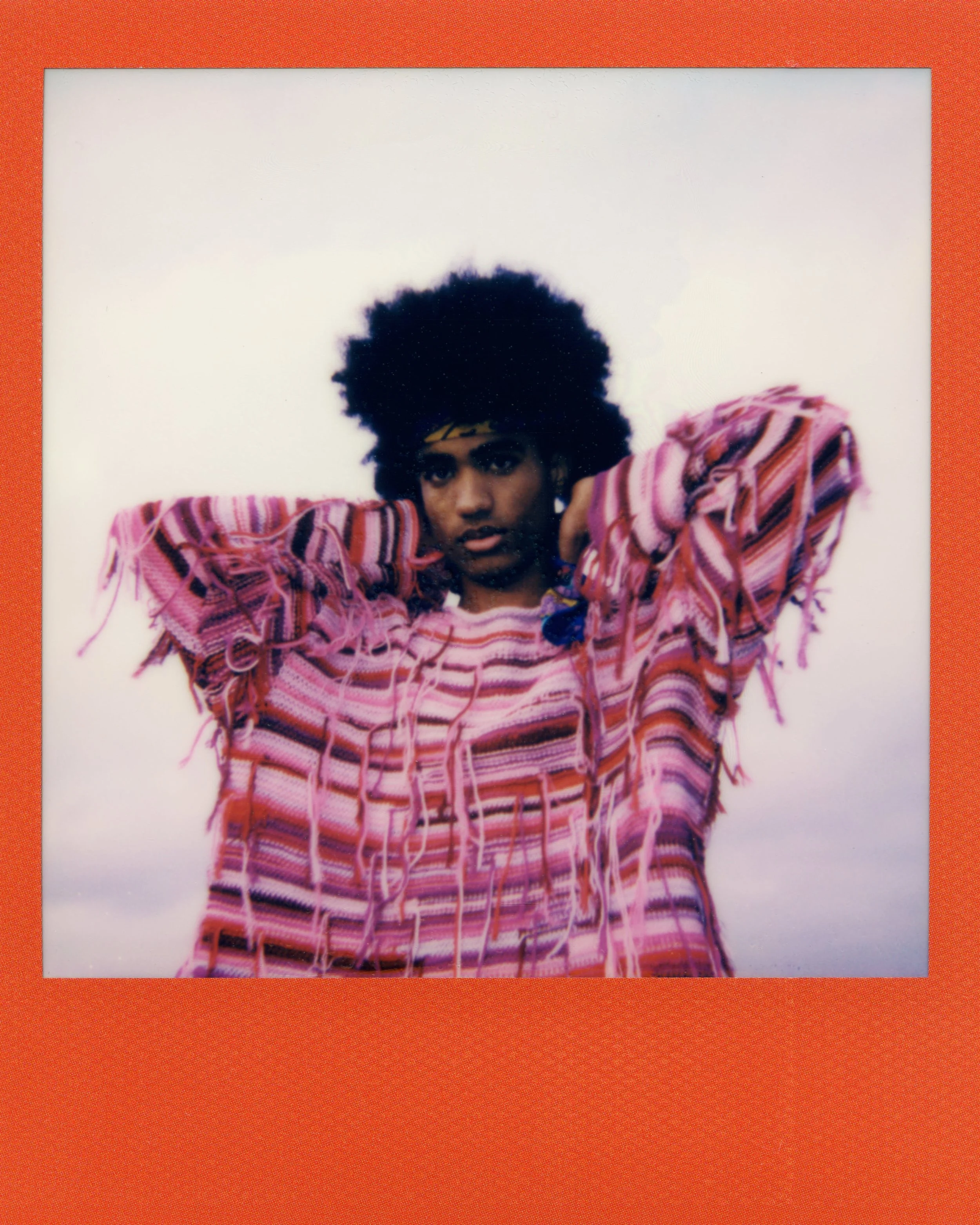 A woman with curly black hair wearing a striped pink, purple, and red sweater, holding her hands up behind her head, looking directly at the camera. The photo has a white background with an orange border.