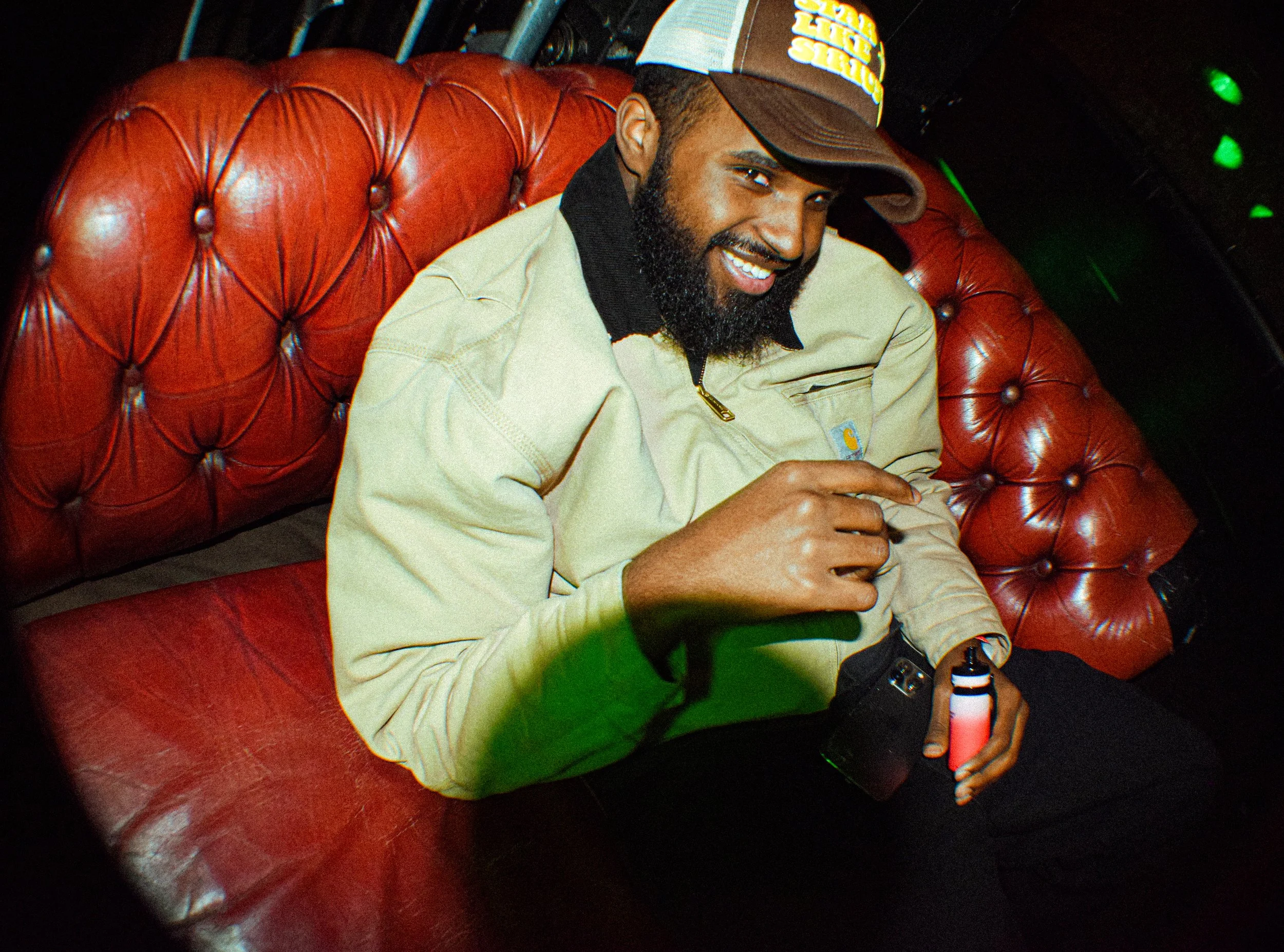 A smiling man with a beard wearing a baseball cap, beige jacket, and black shirt, sitting on a red leather tufted couch, holding a beer bottle in a dimly lit room with green lights.