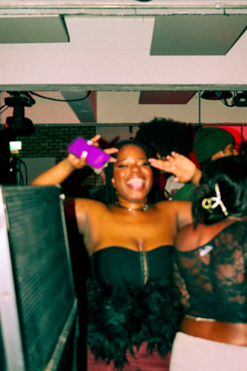 A woman smiling and holding a purple object near her ear at a lively party or event.