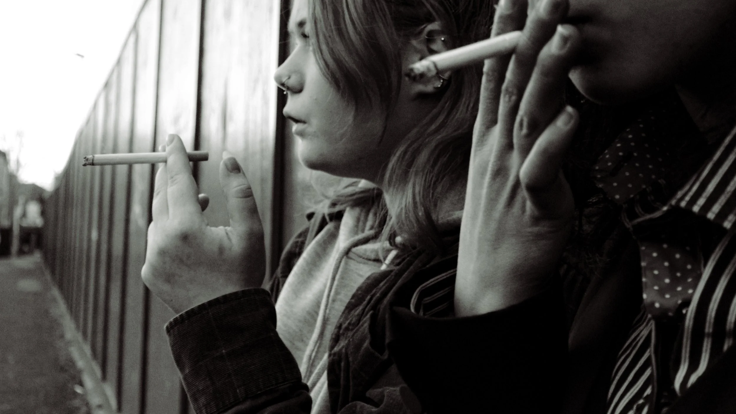 Two young women are sitting side by side outdoors next to a wooden fence, smoking cigarettes.