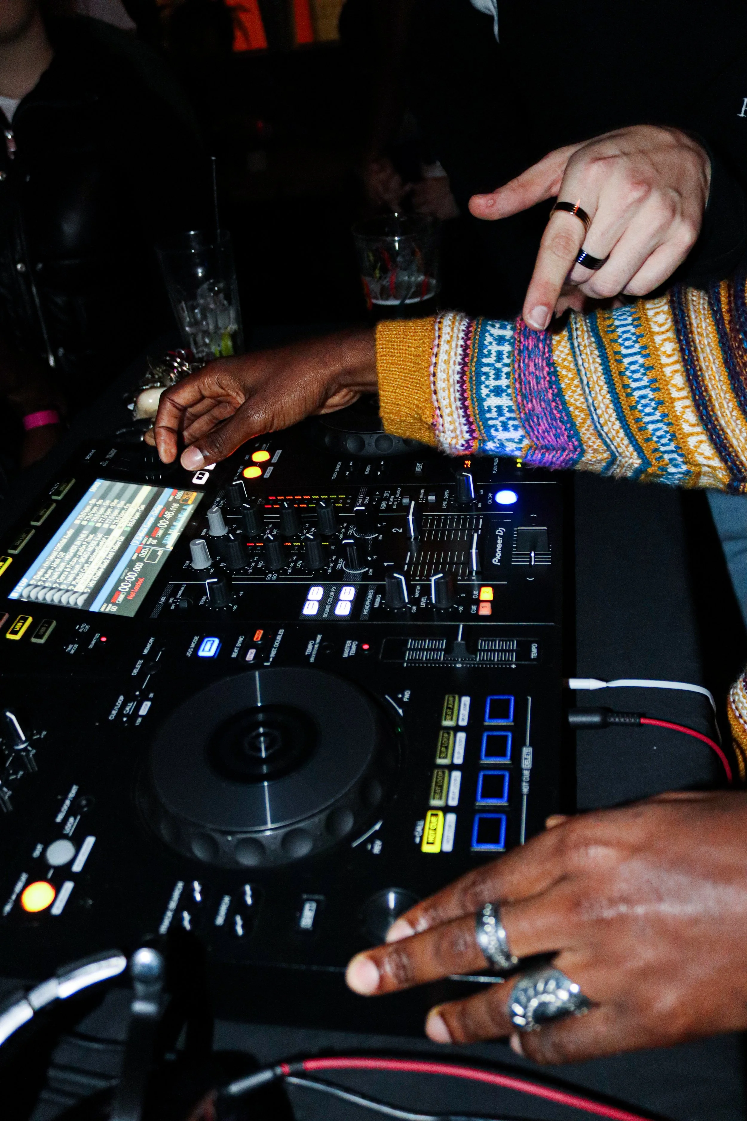Two people, one with dark skin and one with light skin, are using DJ equipment with a mixer and turntable. The dark-skinned person is adjusting knobs, and the light-skinned person is pointing at the equipment. Both are wearing rings and rings are vis