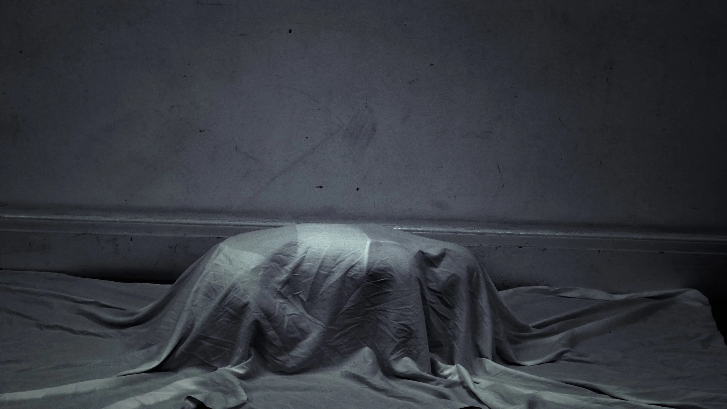 A person covered with a white sheet lying on a bed in a dimly lit room with a gray wall.