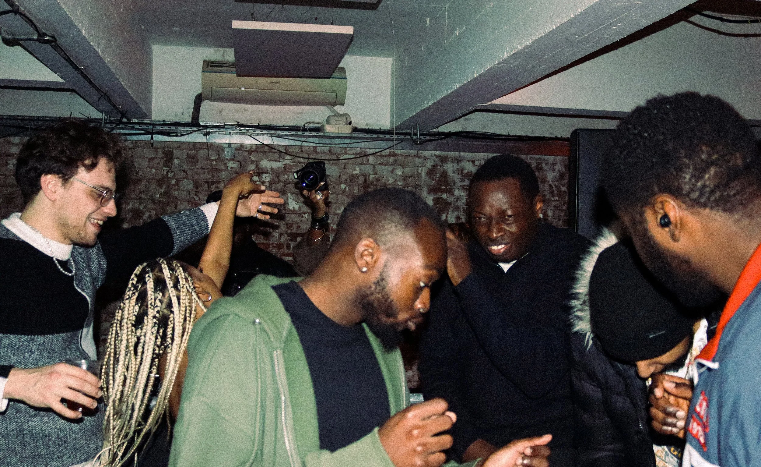 A group of people gathered closely together, smiling and looking down at their phones, in a room with exposed brick walls and a projector on the ceiling.