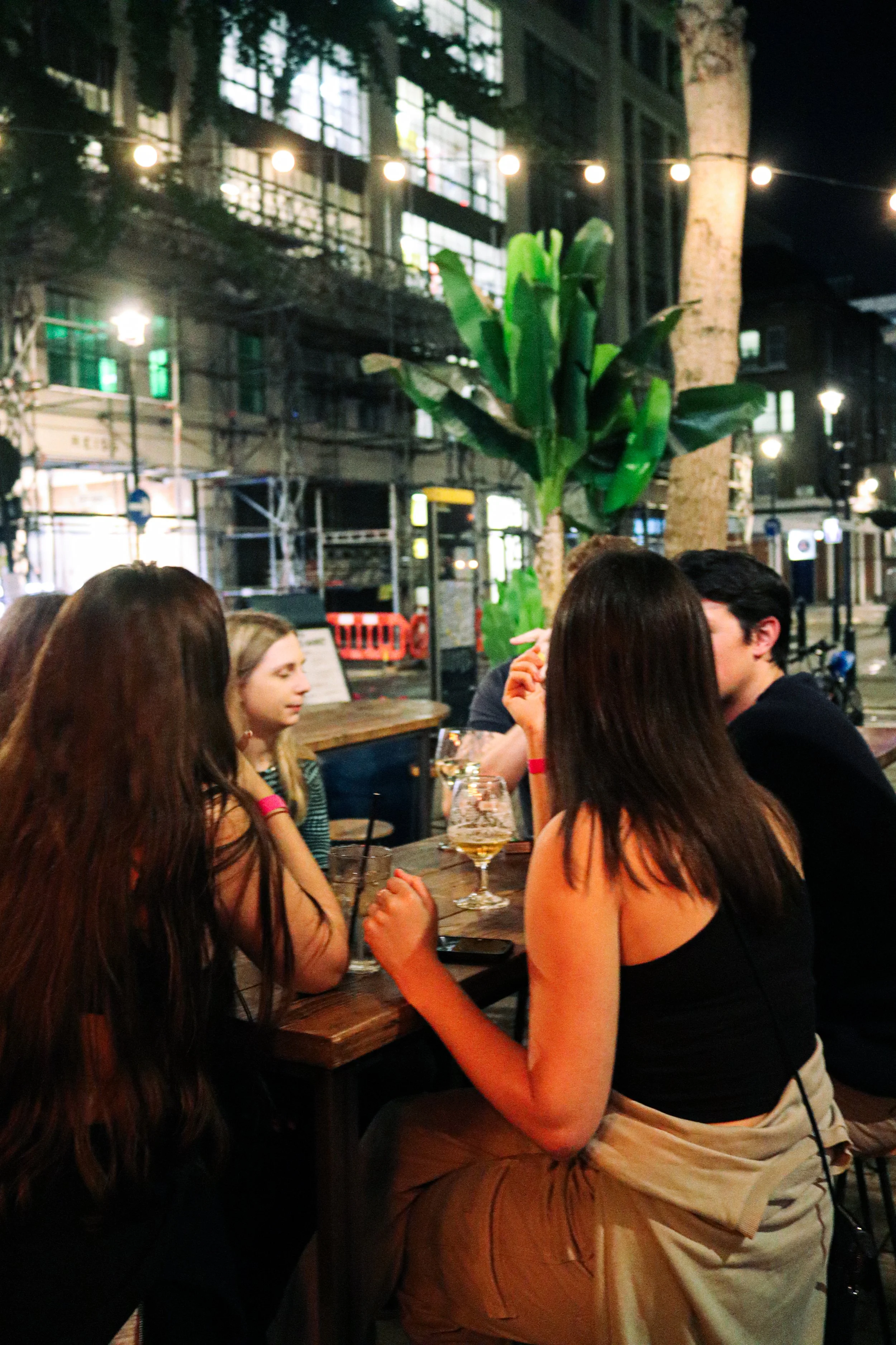 A group of people sitting around a table outdoors at night, engaged in conversation. There are drinks on the table, and the setting includes string lights and a large plant in the background.
