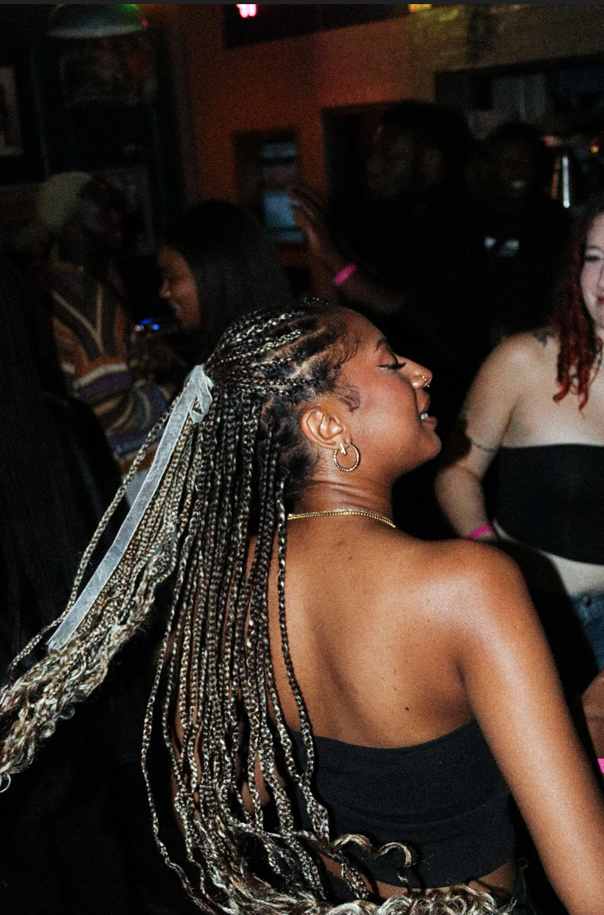 A woman with braided hair and hoop earrings is enjoying herself at a crowded party, with people dancing and smiling in the dimly lit background.