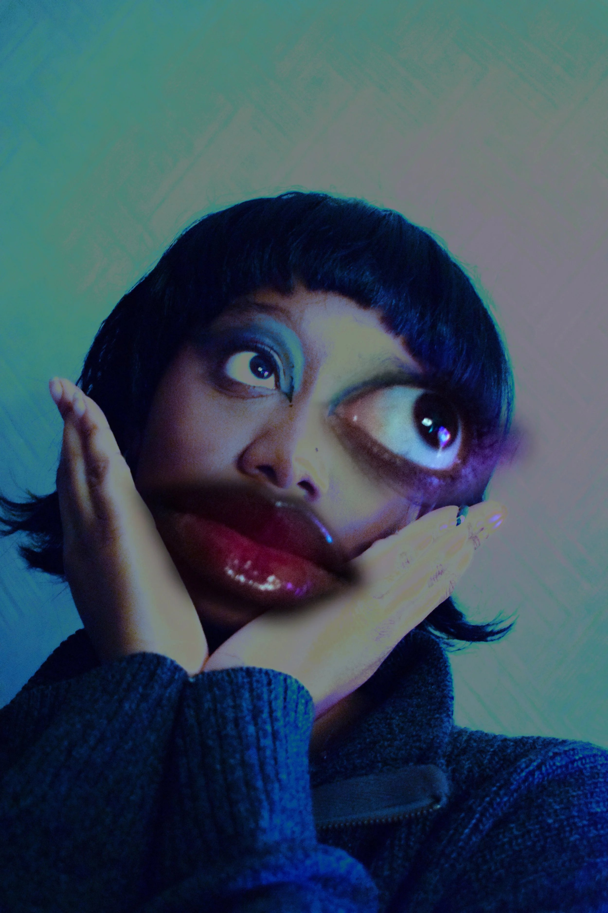 Close-up of a woman with short black hair and expressive eyes, holding her face with hands. She is wearing a dark blue or black sweater, and the background is plain with muted colors.