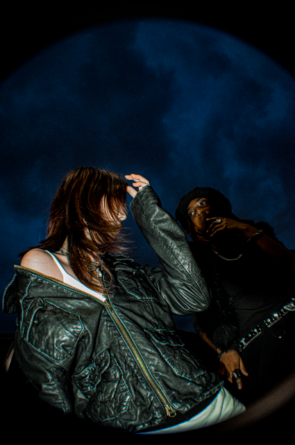 Two women standing outdoors at night, with a large, bright full moon in the background. One woman has brown hair and is wearing a leather jacket, while the other woman has dark hair and is wearing a black outfit.