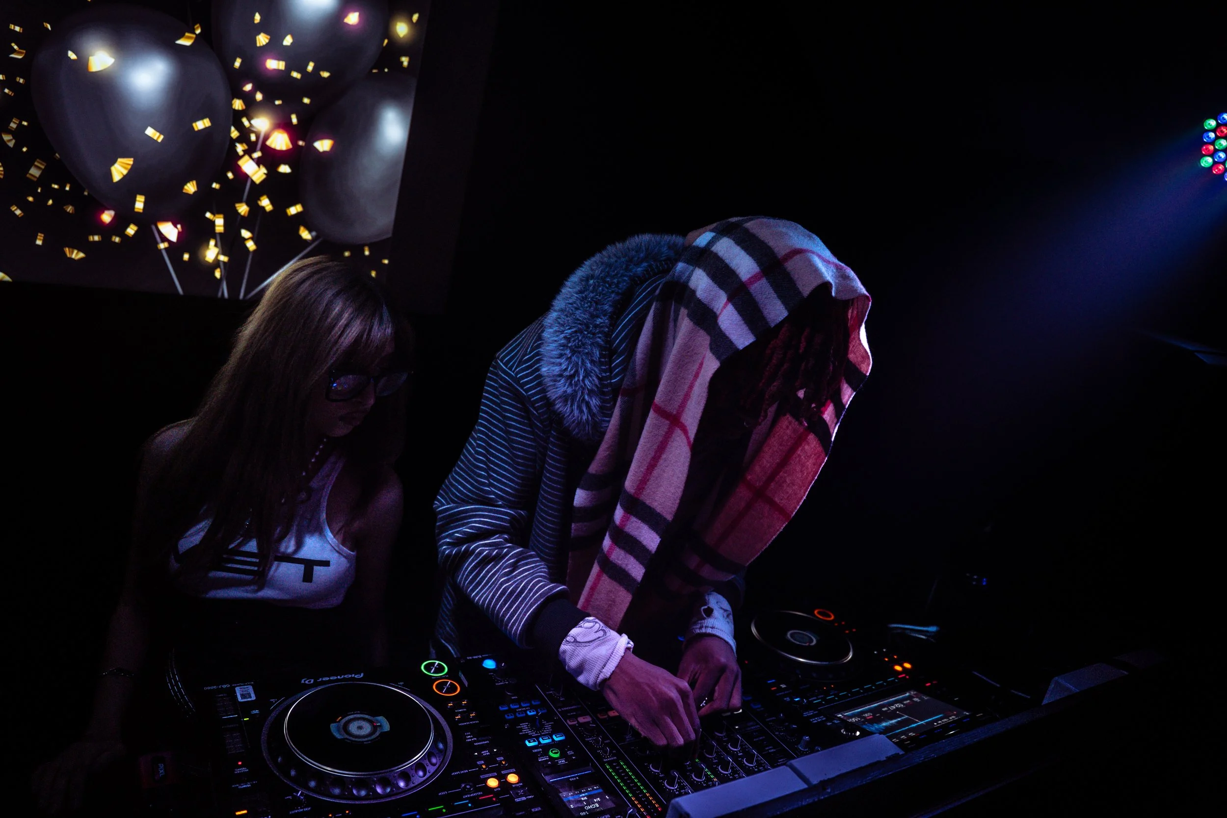 Two people, one with long hair and glasses and another with a striped shirt and a checkered towel over the head, are DJing at a dark party venue with colorful lights.