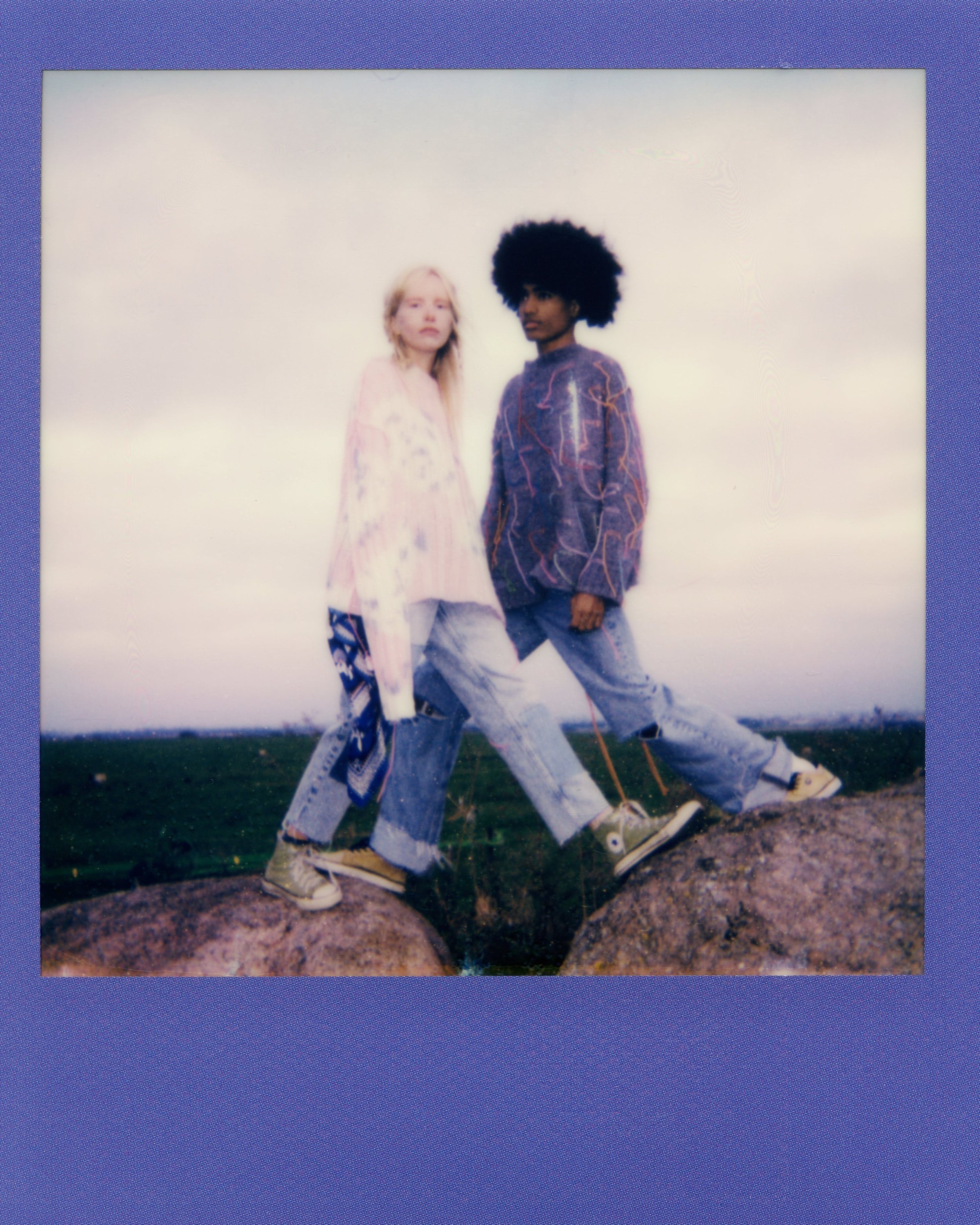 Two young women standing outdoors on rocky ground with cloudy sky in background. One woman has blonde hair and wears a light-colored dress, the other has dark curly hair and wears a purple sweatshirt. Both are wearing jeans and sneakers.
