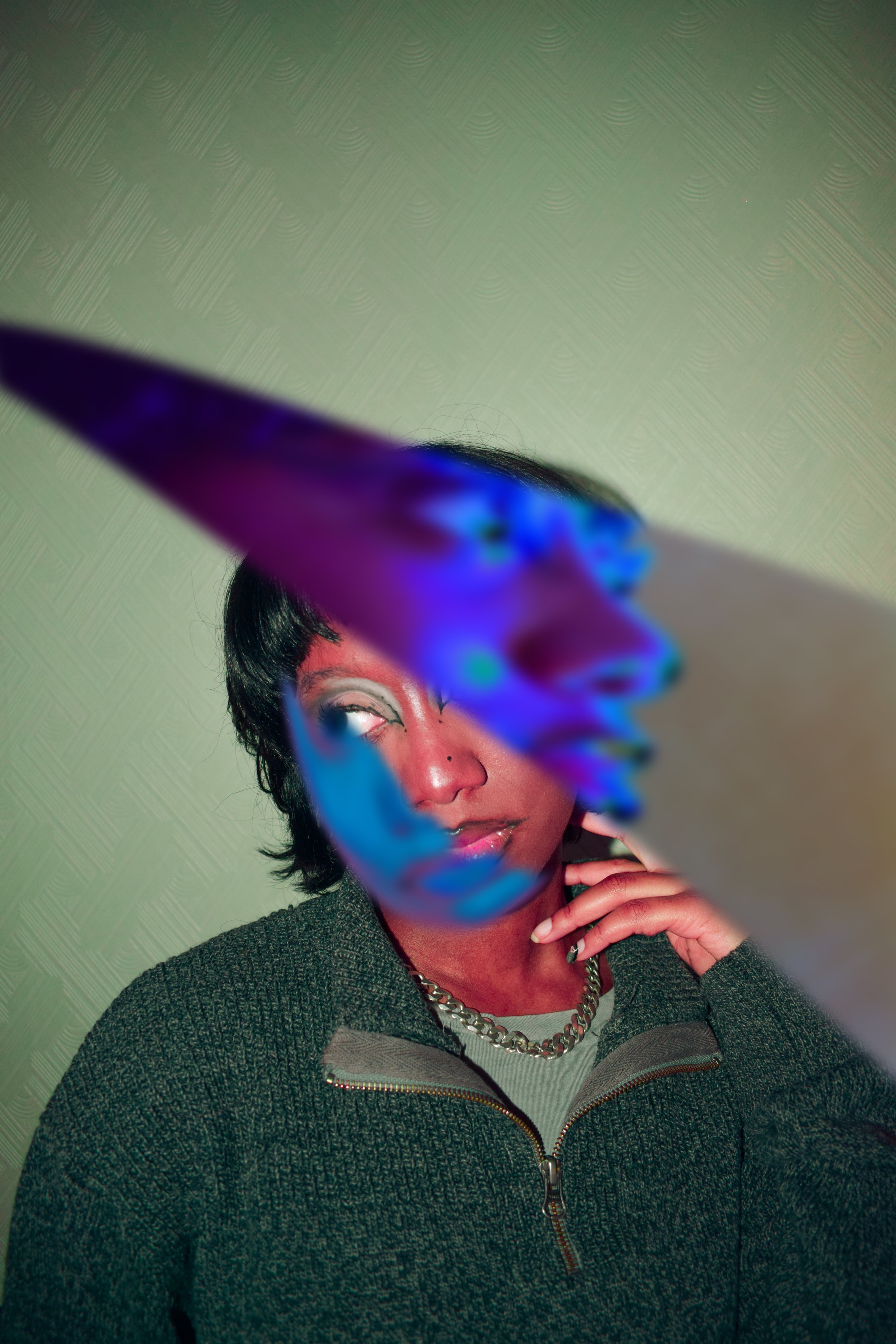 A person with dark hair, wearing a gray textured jacket and a chain necklace, holding a colorful abstract object in front of their face, covering part of it, with a plain green background.