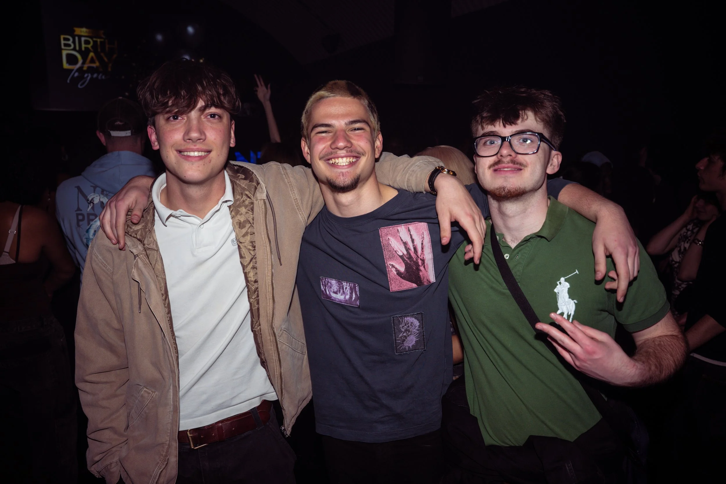 Three young men smiling and posing together at a party or nightclub with a dark background and other people in the distance.