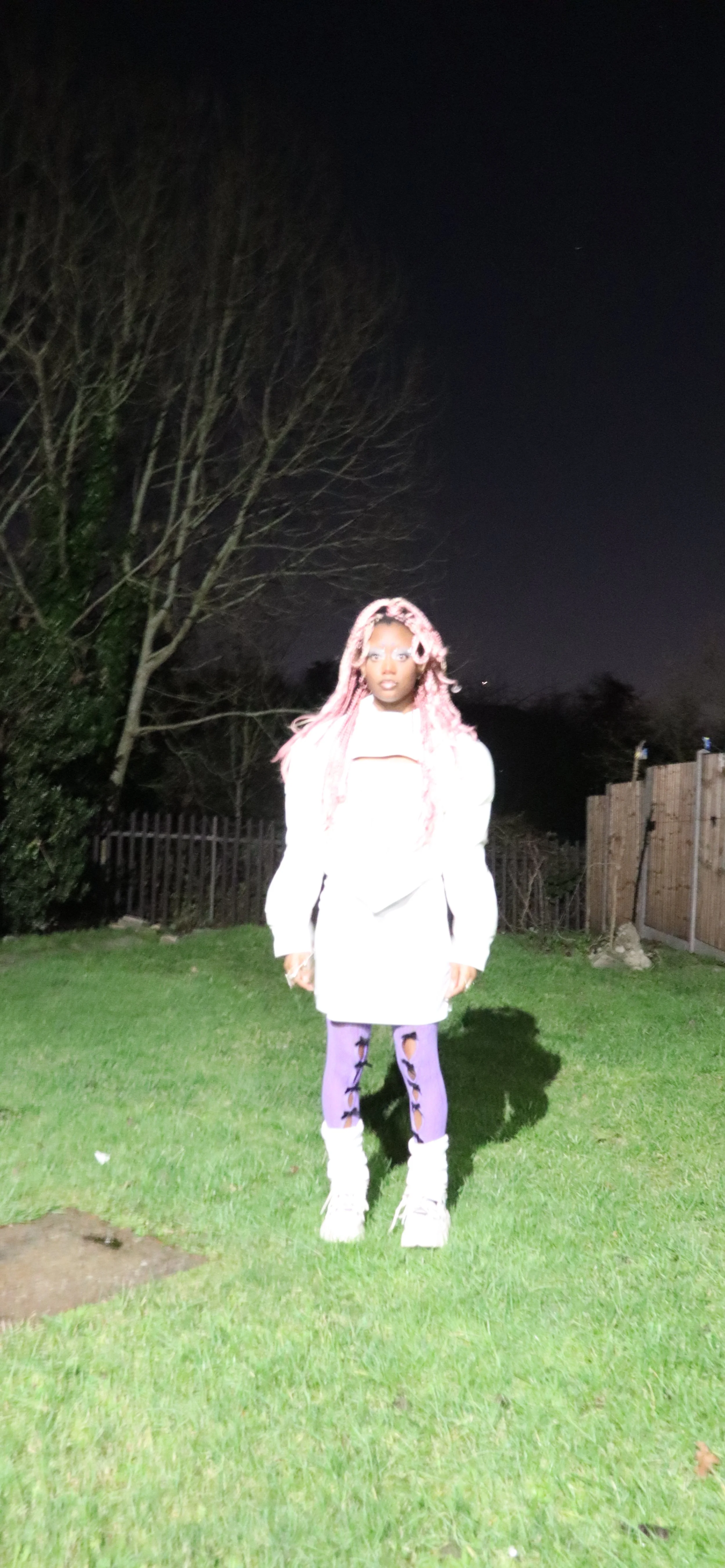 A person with pink braided hair standing on a grassy lawn at night, wearing a white coat, purple leggings with black patterns, and white shoes, with a dark night sky and trees in the background.