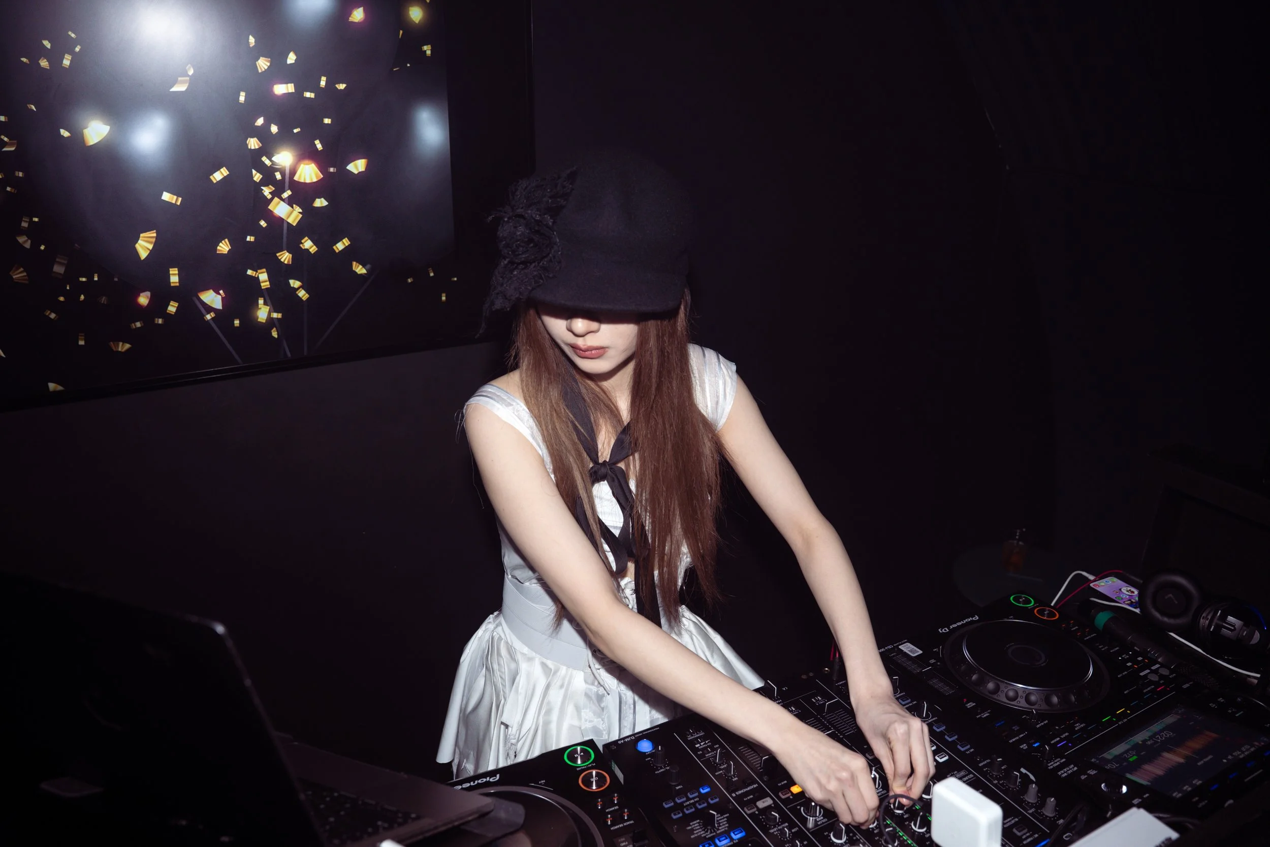 A woman with long reddish hair wearing a black hat and a white dress is DJing at a nightclub or party. She is focused on the DJ equipment in front of her, surrounded by dark walls with a visual display of golden confetti and lights on a screen behind her.
