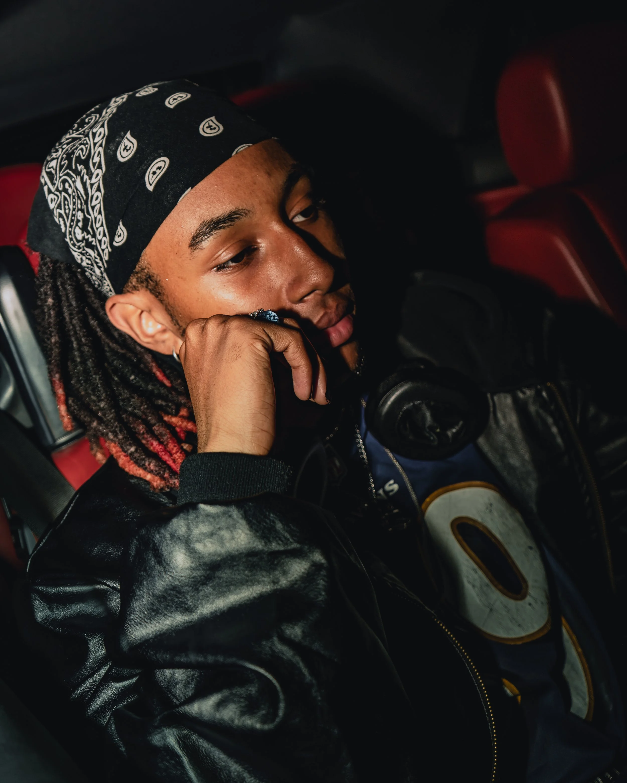 A young person wearing a black bandana with white patterns, holding a pair of headphones and sitting in a vehicle with red interior seats.