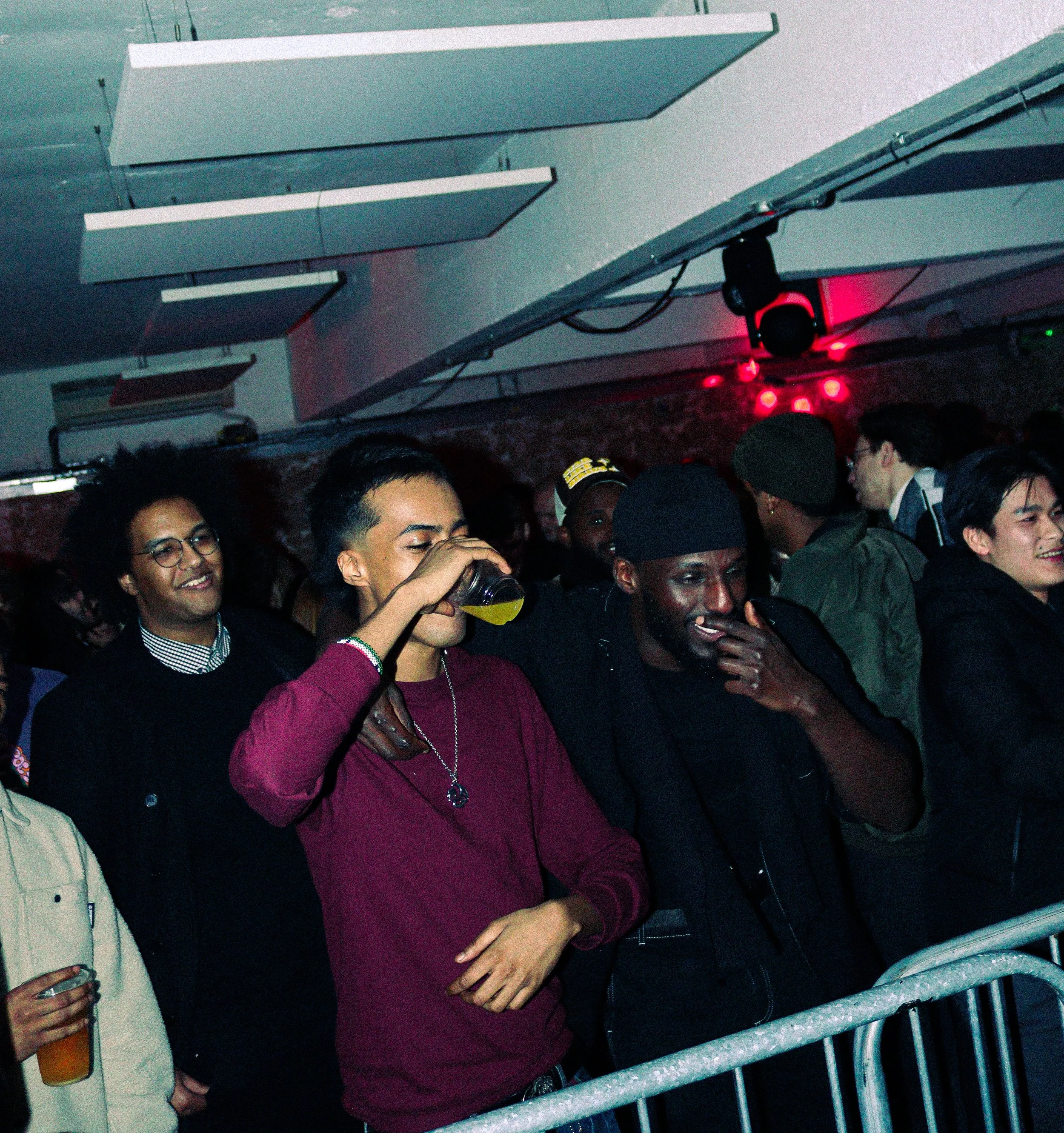 A group of young people at a concert or club, some smiling and enjoying drinks, with red and blue lighting and a metallic railing in the foreground.