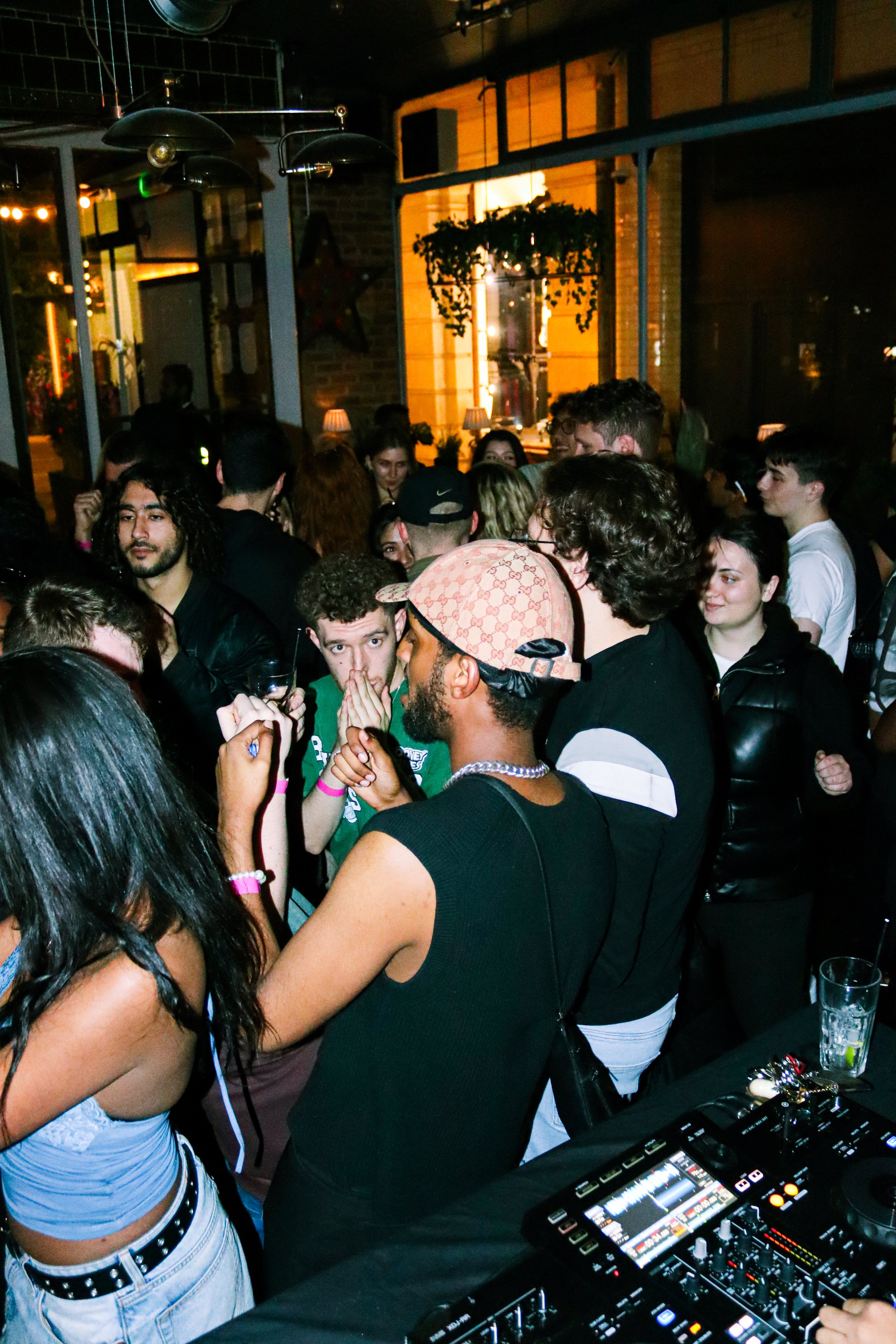 A crowded indoor party with people dancing and socializing, a DJ deck with equipment in the foreground, and warm lighting inside a modern venue with large windows and decorative elements.