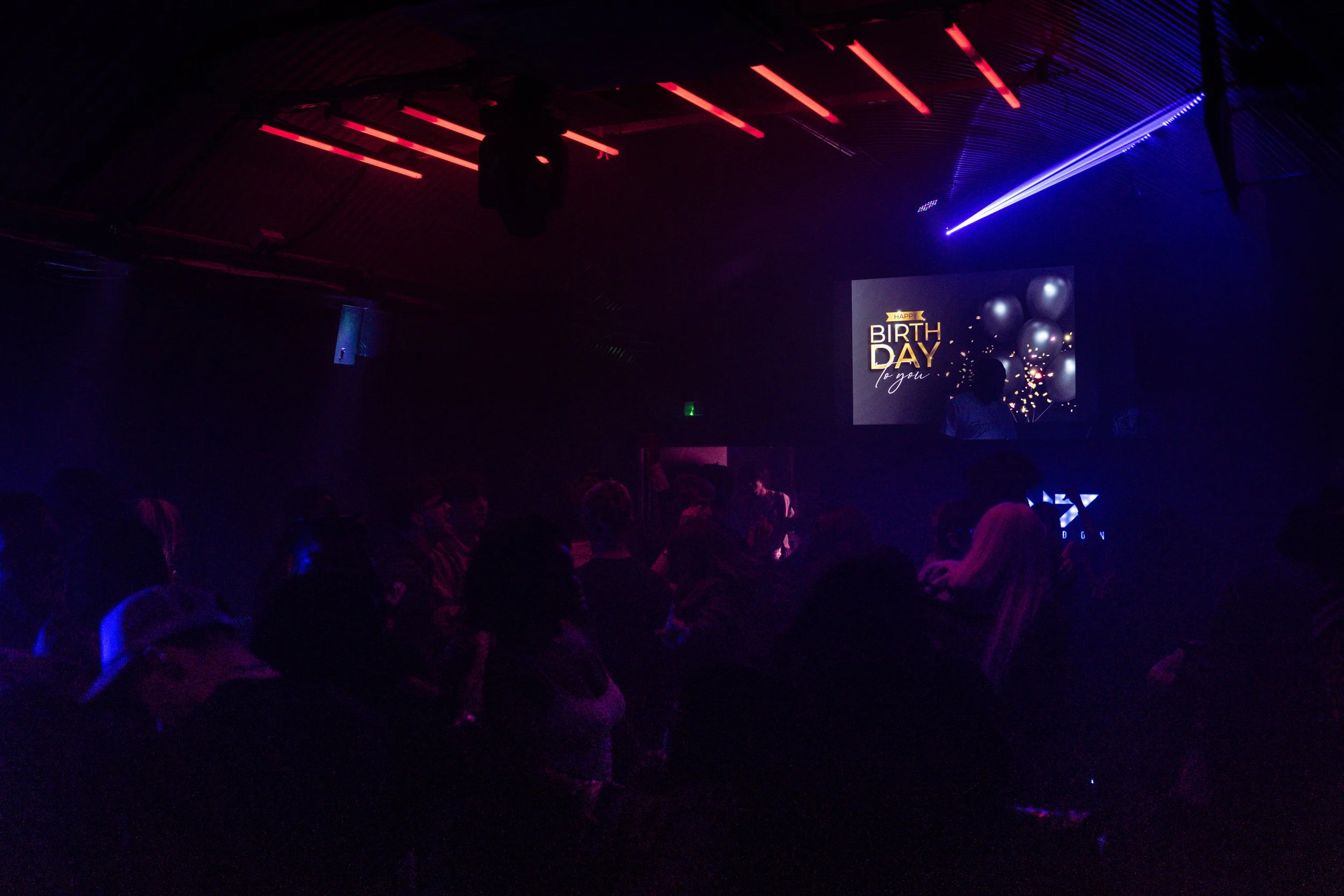 A dark room filled with people and a large screen displaying a birthday message with balloons and confetti.