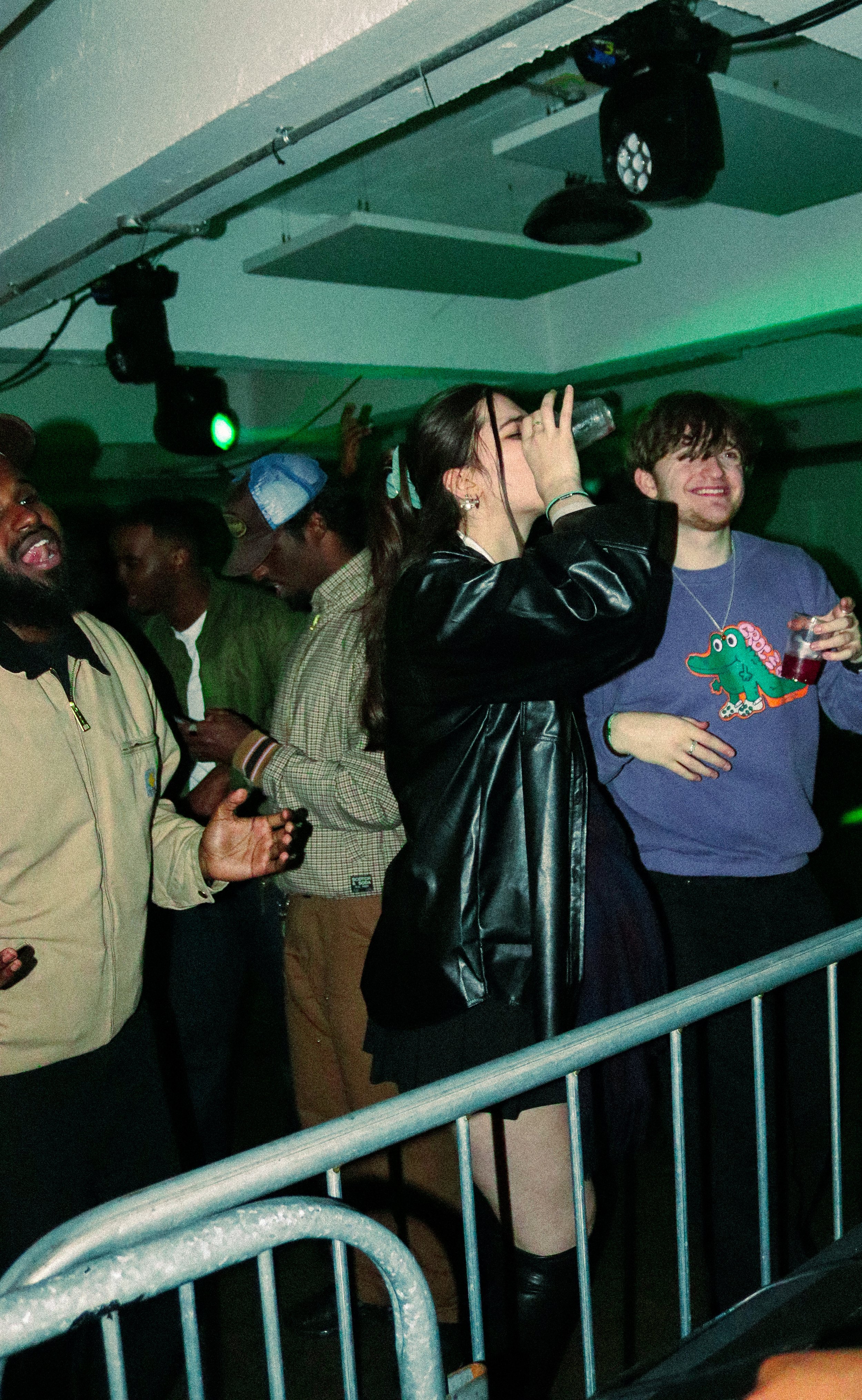 People dancing and enjoying music at a concert or party, some standing behind a metal railing, with green lighting overhead.