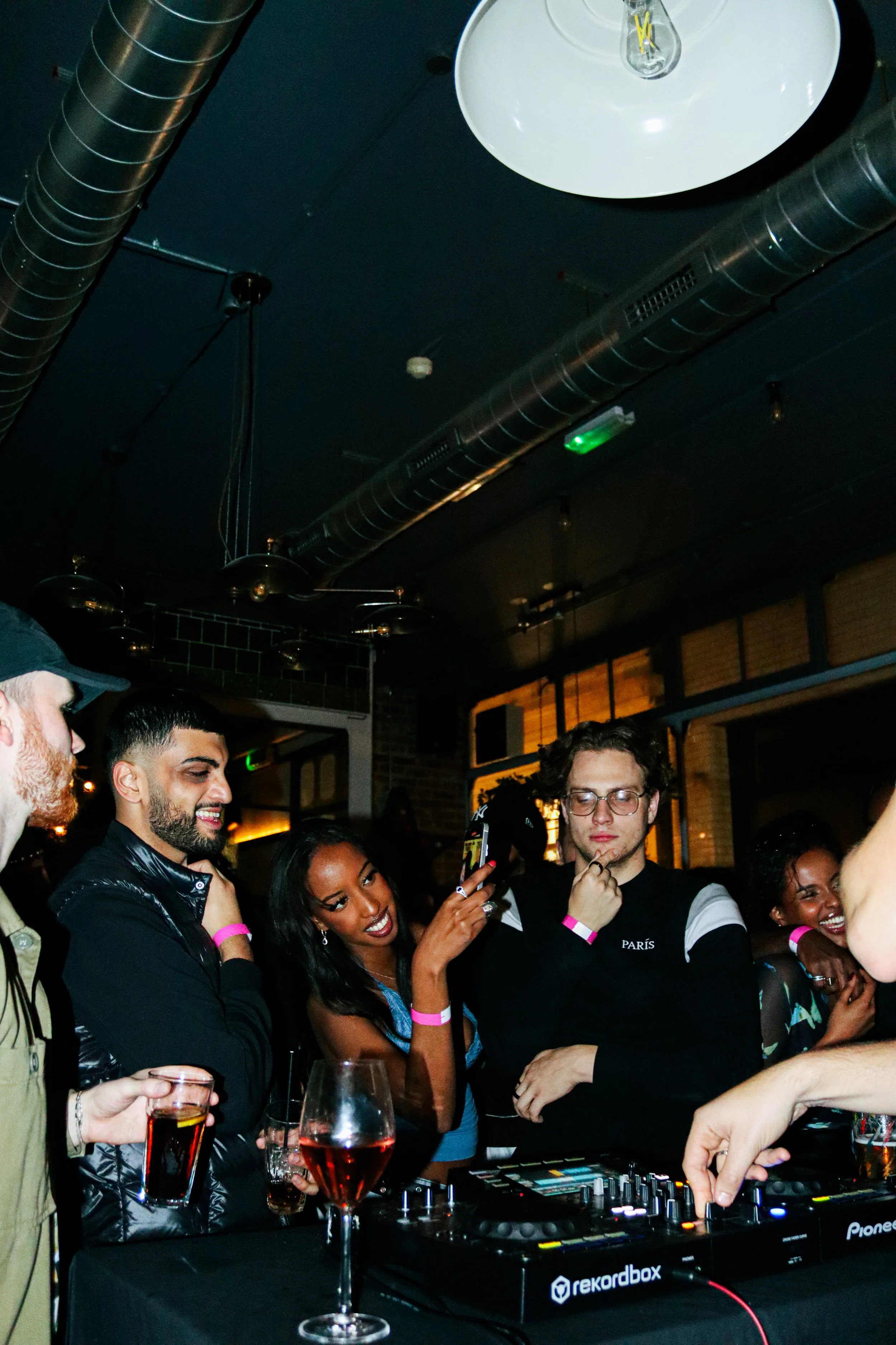 A group of people at a DJ booth in a dimly lit bar or nightclub. Some are holding drinks and smiling, with a DJ controller in front of them. The background features industrial-style ductwork on the ceiling and warm lighting.