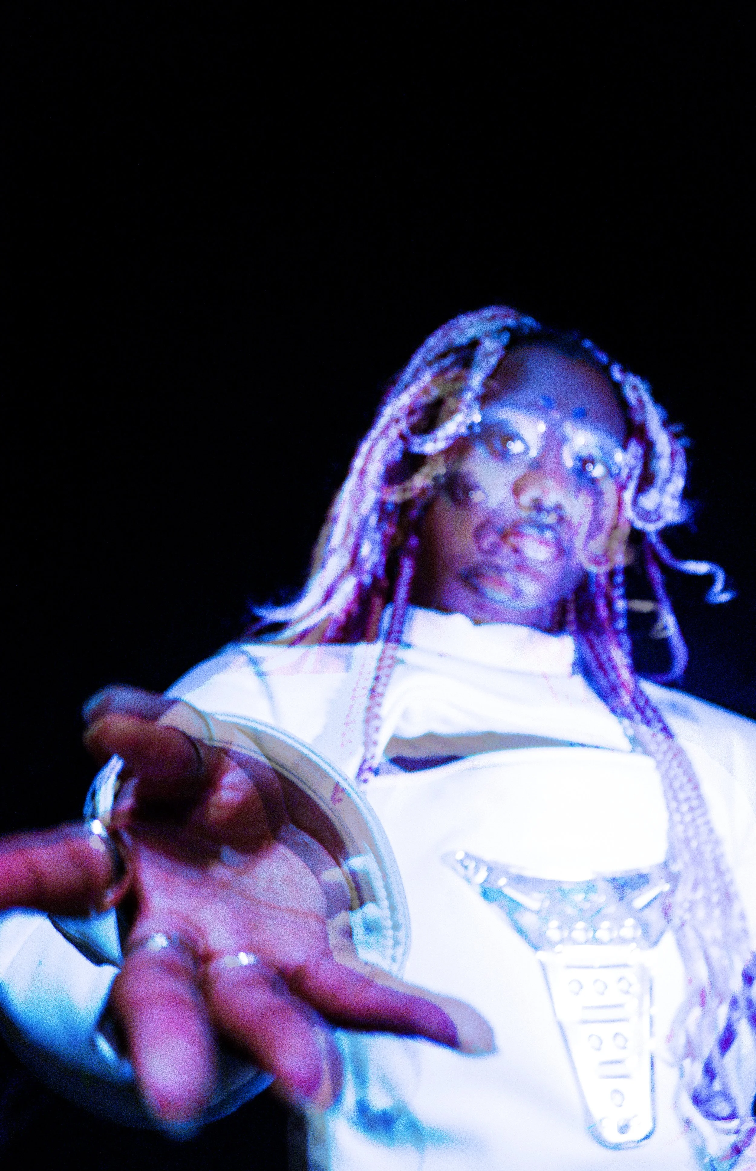 Close-up of a person in a white outfit with a microphone, reaching out with their hand, head with colorful braids and face with makeup, against a dark background.