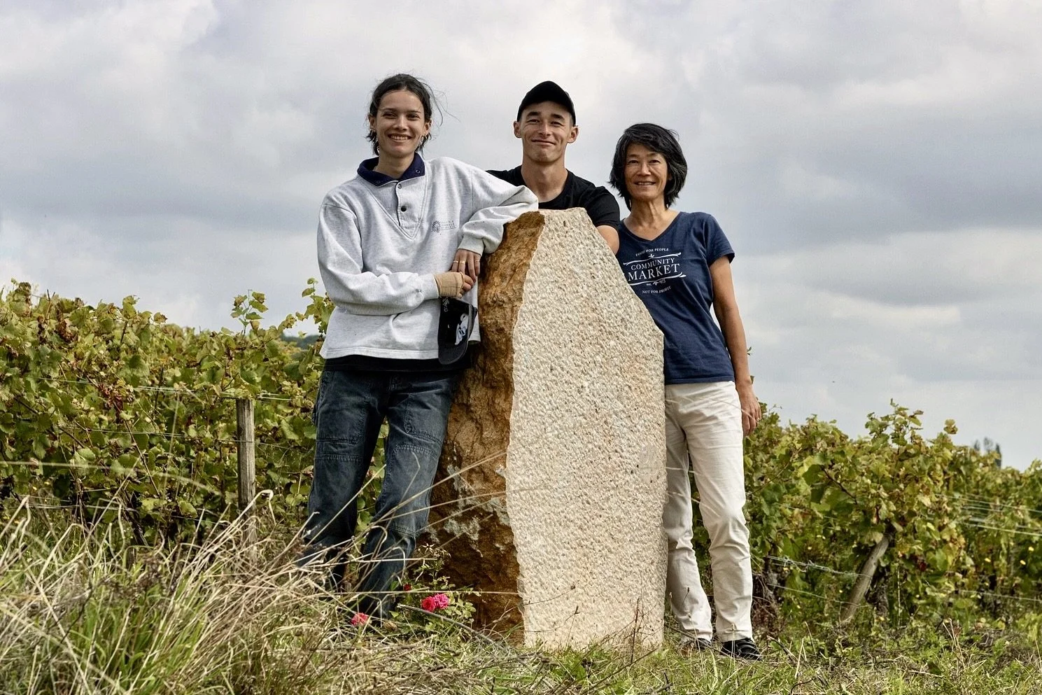 Trois personnes debout dans un vignoble, souriantes, s'appuyant sur une pierre en pierre, sous un ciel nuageux.