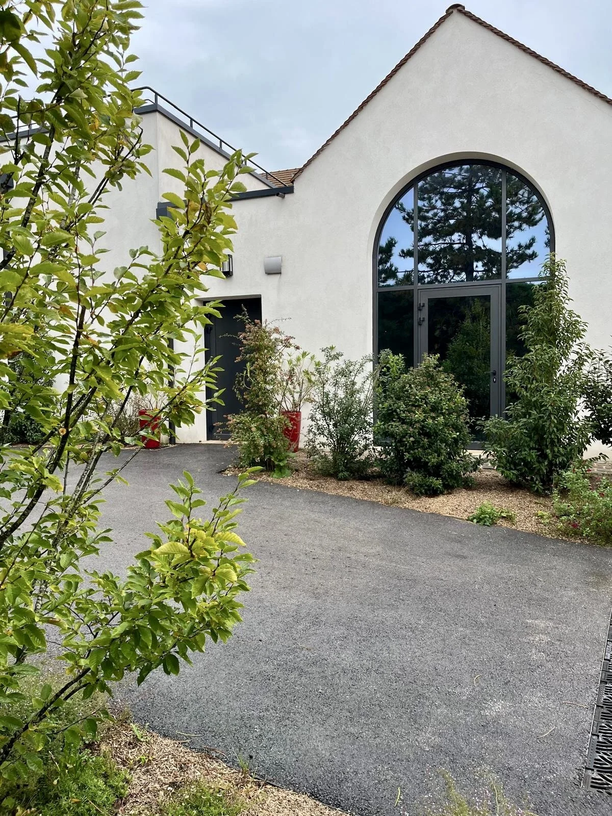 Vue d'une maison moderne avec une grande fenêtre arrondie, entourée de plantes et de buissons, avec une allée en asphalte devant.