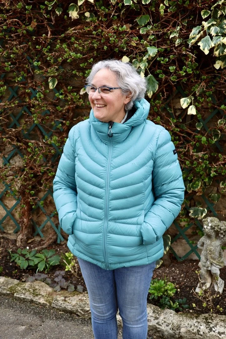 Femme souriante portant une veste bleue, debout devant une haie verte, en extérieur.