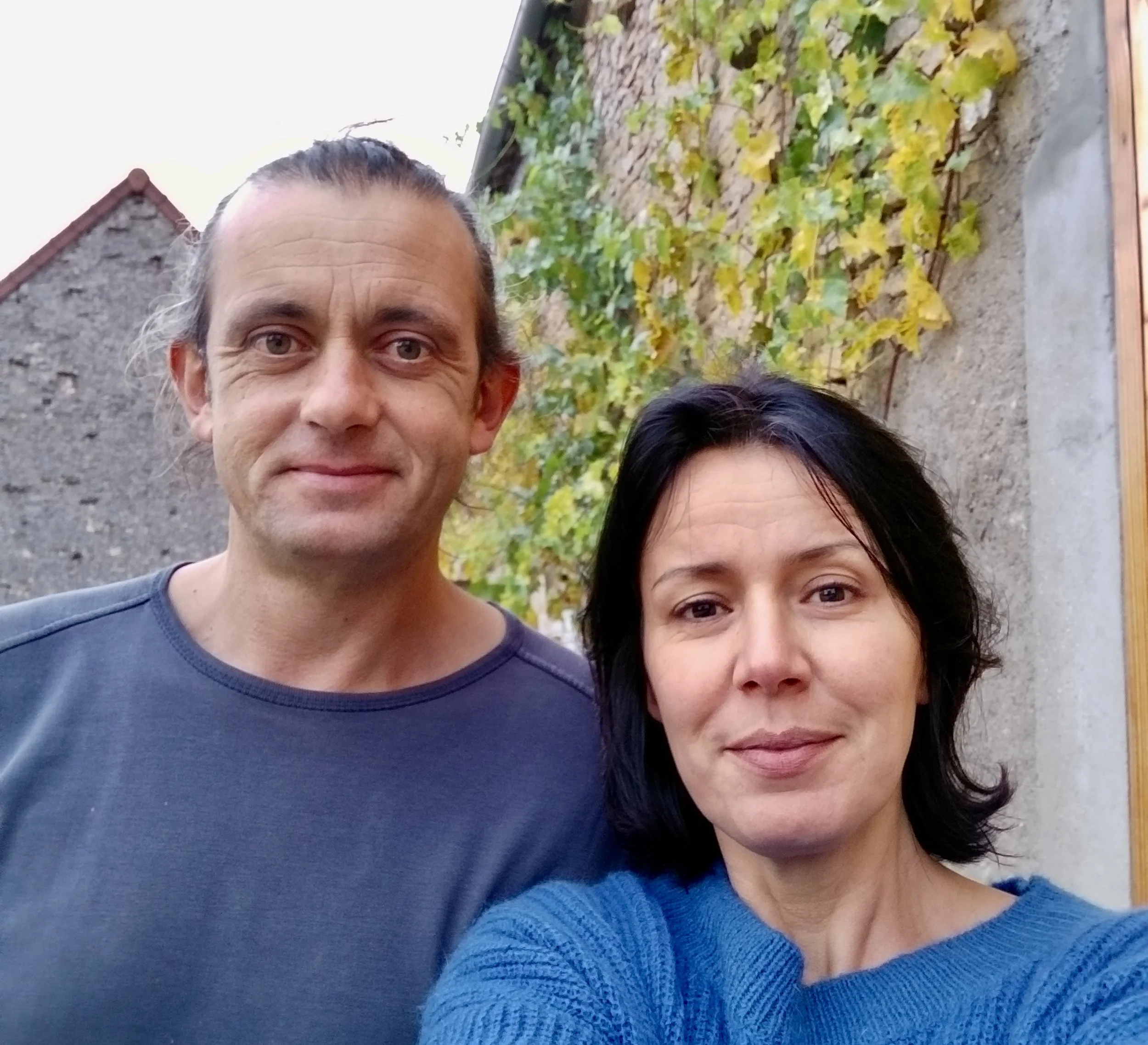 Un homme et une femme prennent un selfie à l'extérieur, avec des murs en pierre et du feuillage pourpres en arrière-plan.
