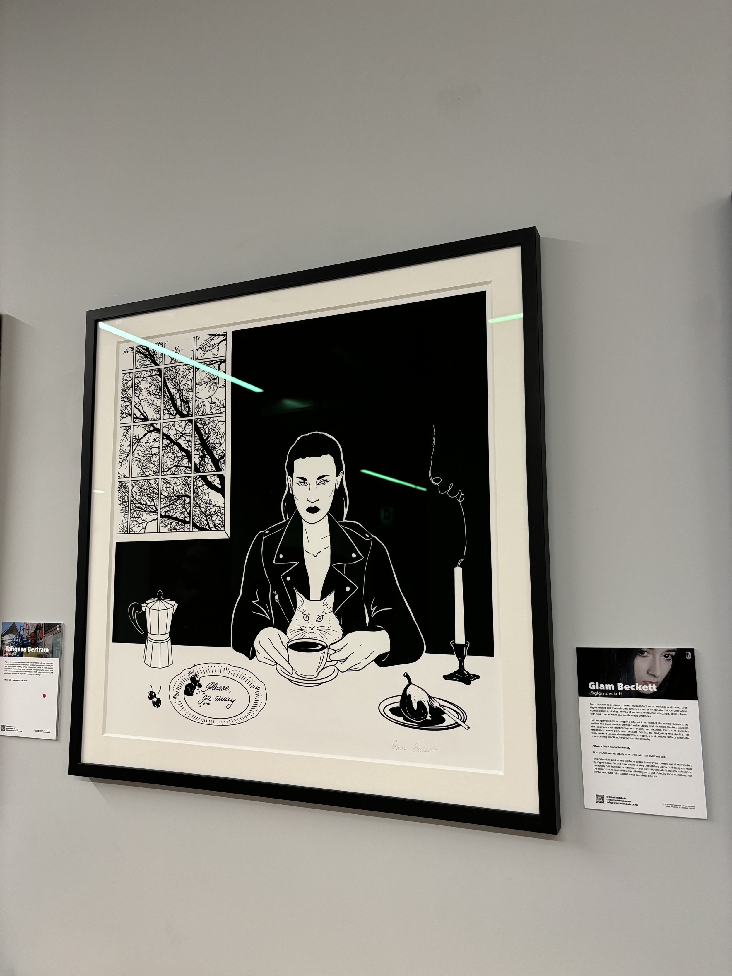 Glam Beckett Exhibition London. Two framed artworks on a wall above a wooden table. The left piece is a black and white illustration of a woman with a cat, coffee, and a lit candle.