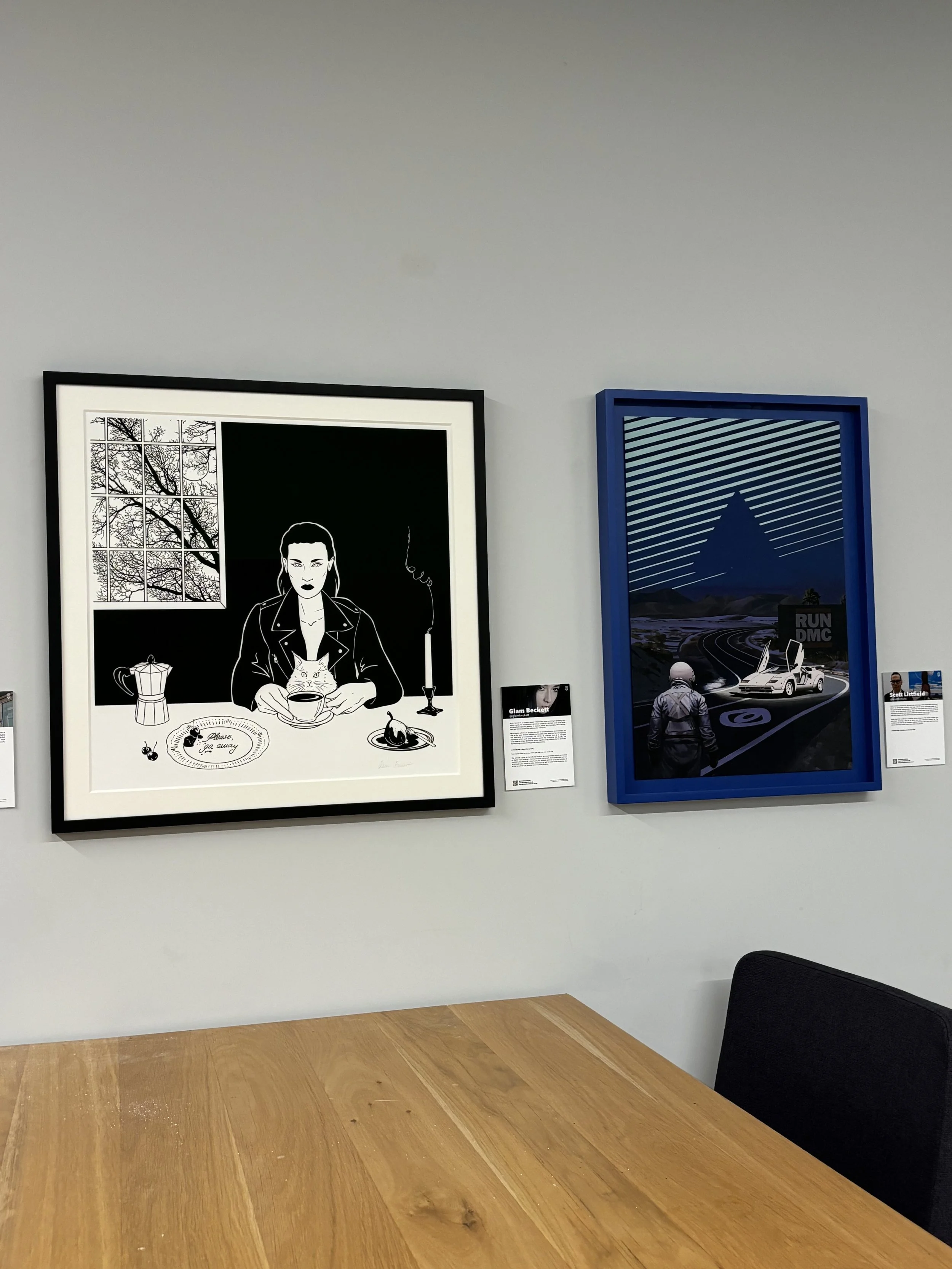 Glam Beckett Exhibition London. Two framed artworks on a wall above a wooden table. The left piece is a black and white illustration of a woman with a cat, coffee, and a lit candle.