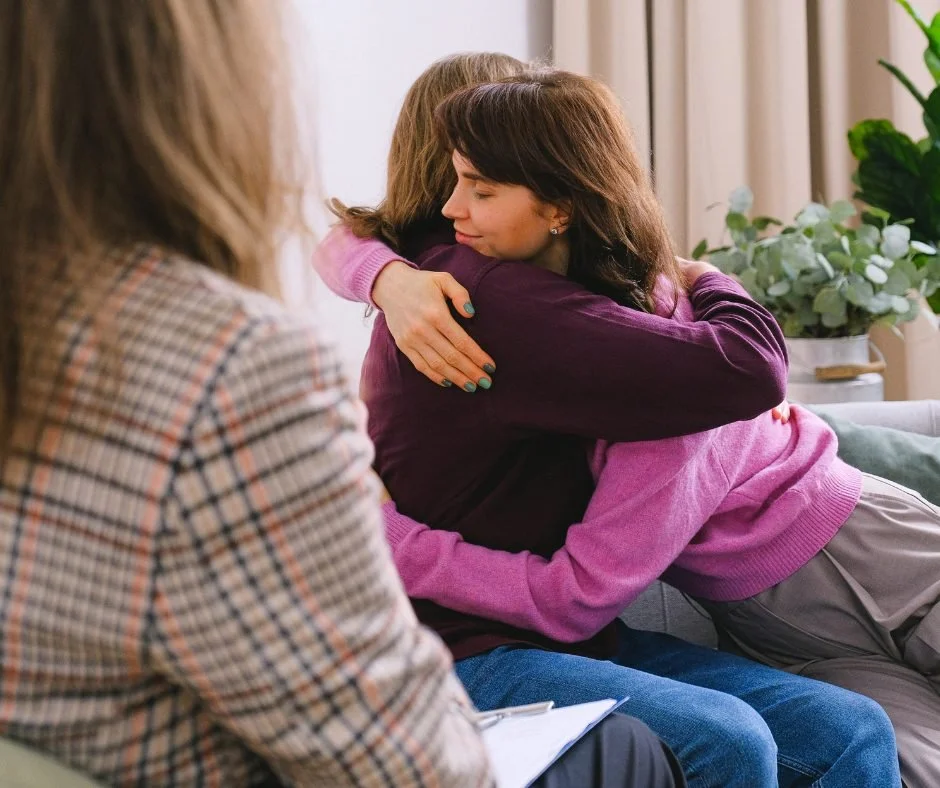 A woman and a girl sharing a hug with another woman sitting nearby, possibly in a counseling or supportive setting.