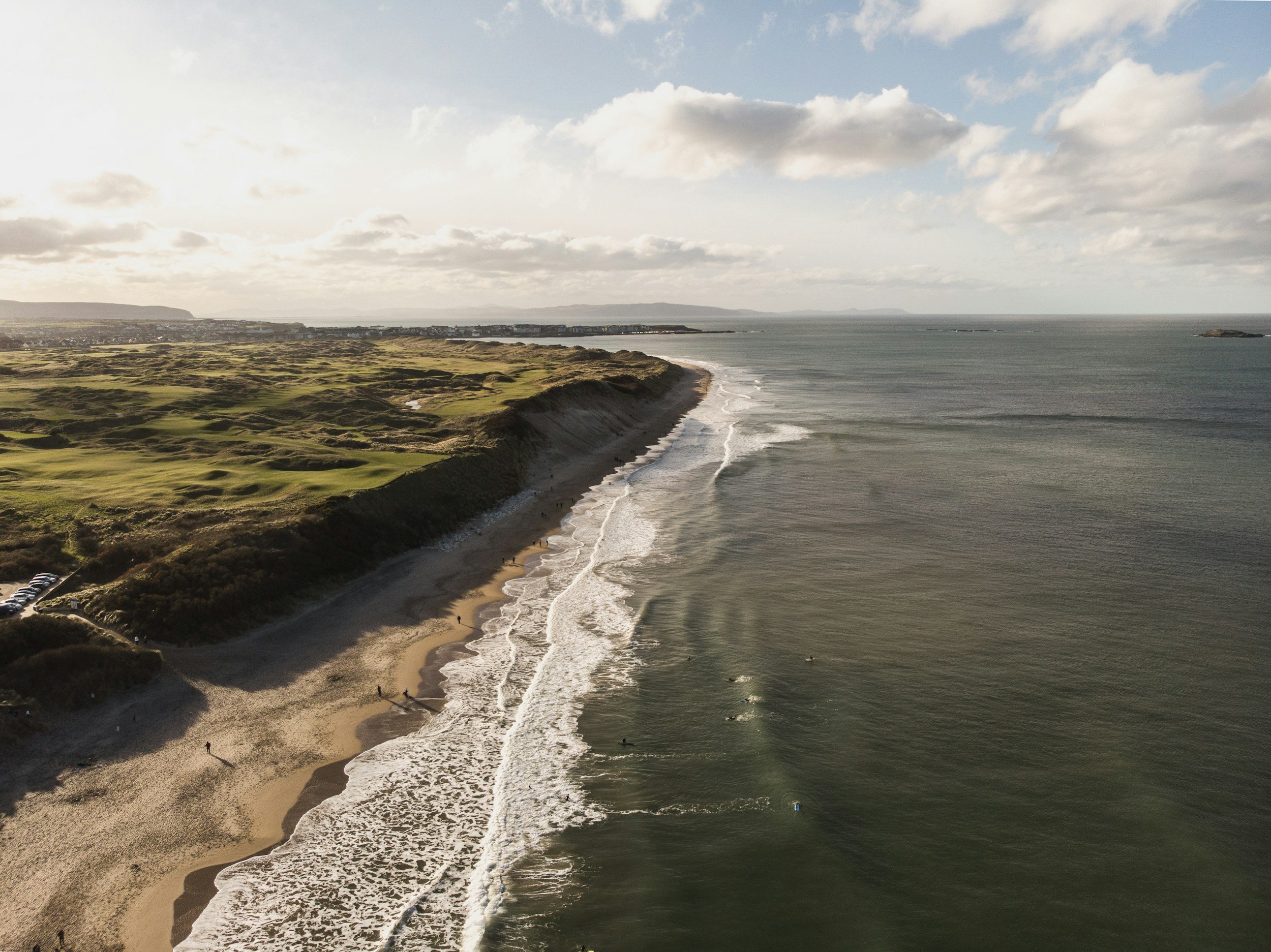 Aerial view of a coastline with sandy beach, waves, and a golf course on green cliffs under a partly cloudy sky.