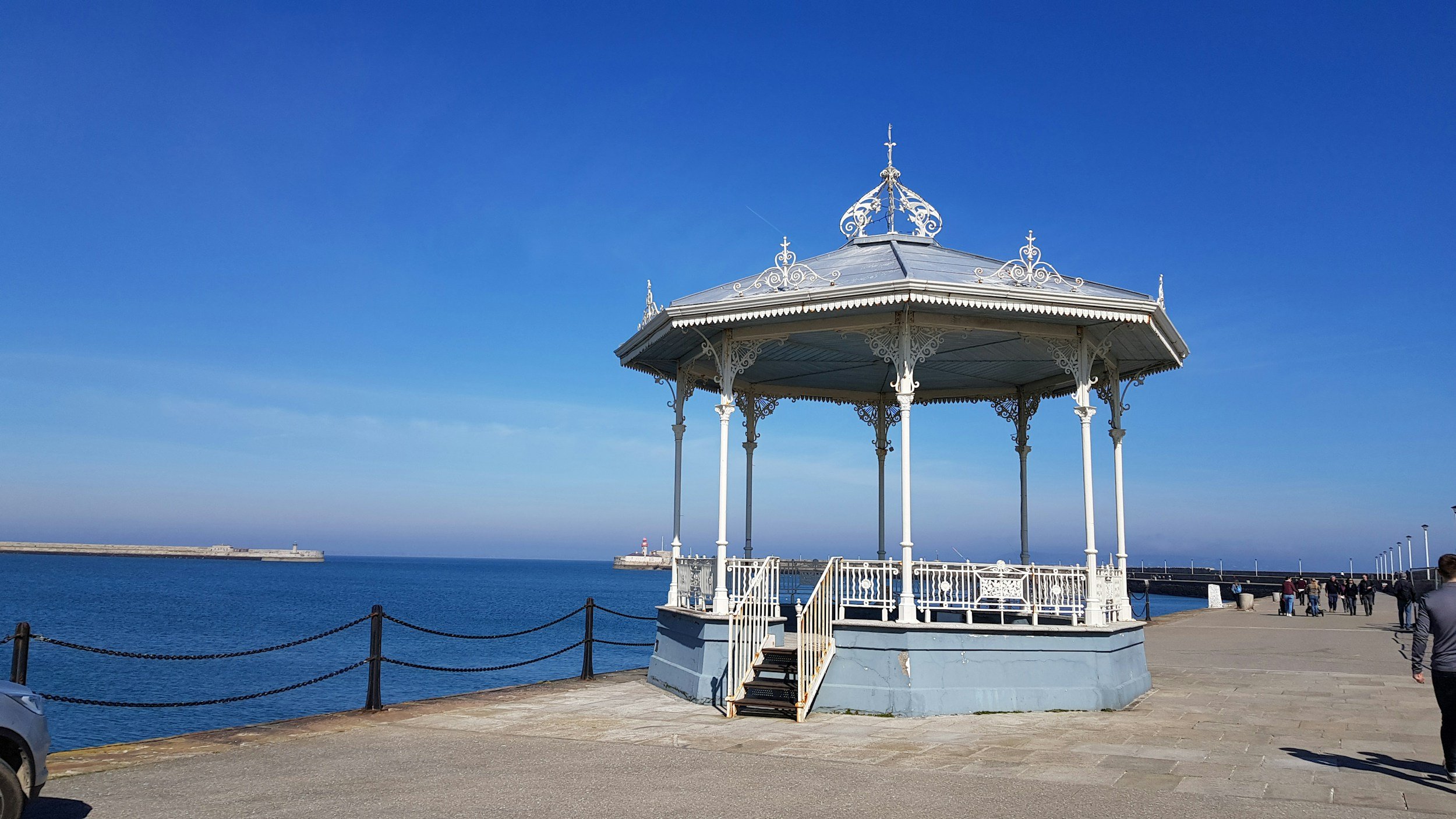 A white, ornate gazebo on a pier by the water under a clear blue sky with a distant lighthouse.