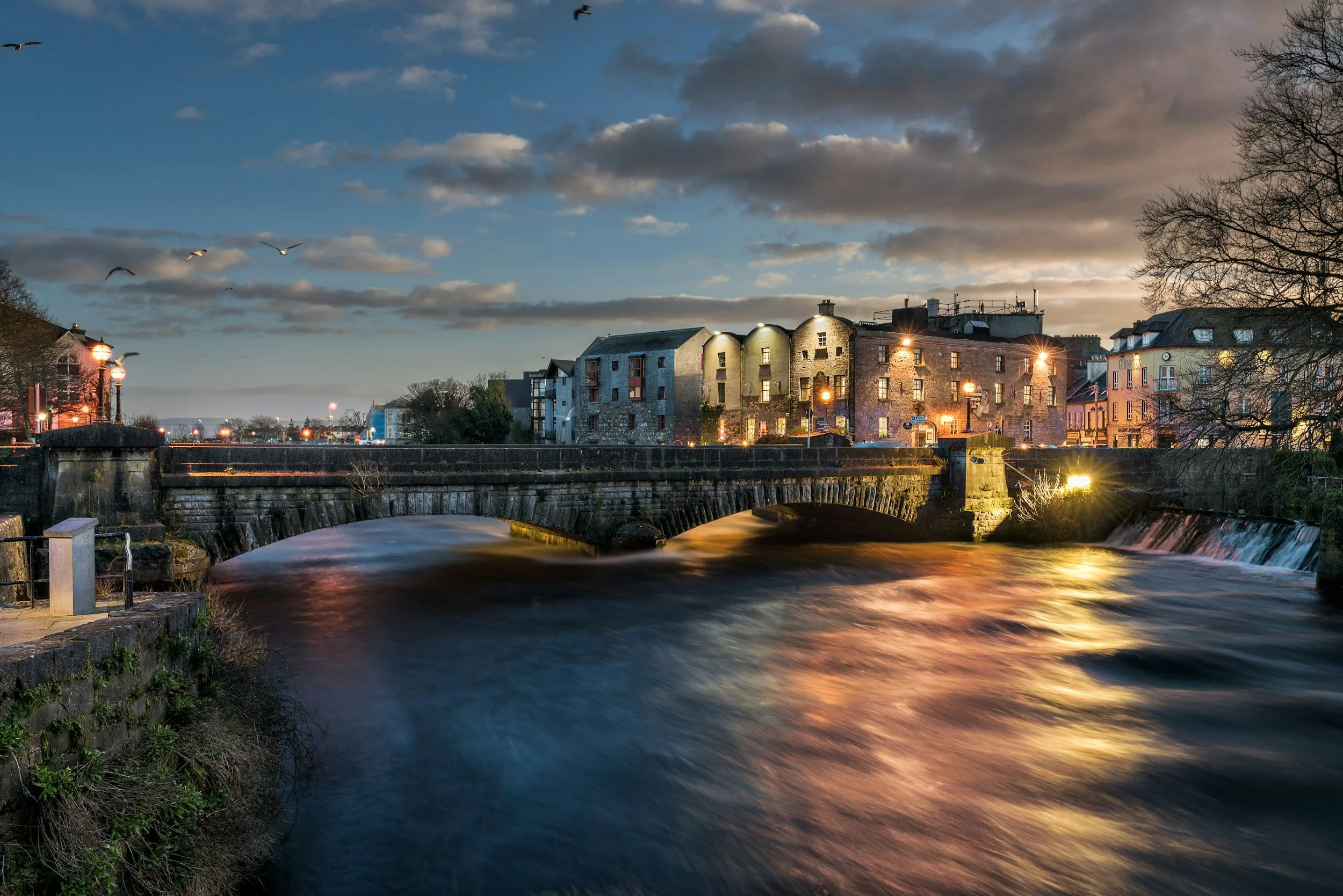 A river flowing under a stone bridge during dusk, with historic buildings lit by streetlights on the riverbank and birds flying in the sky.