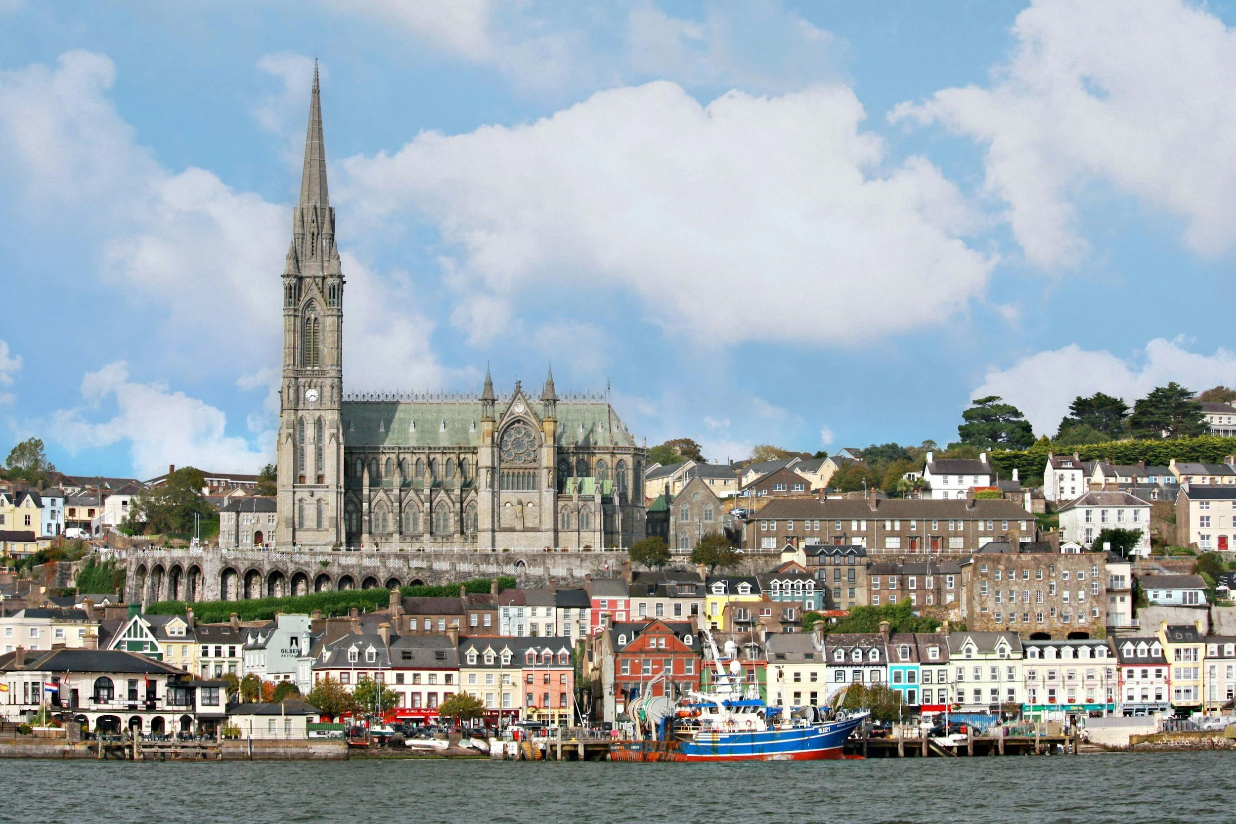 A scenic view of a large Gothic-style church with a tall steeple, surrounded by colorful houses on a hillside, with a harbor and boats in the foreground, under a partly cloudy sky.