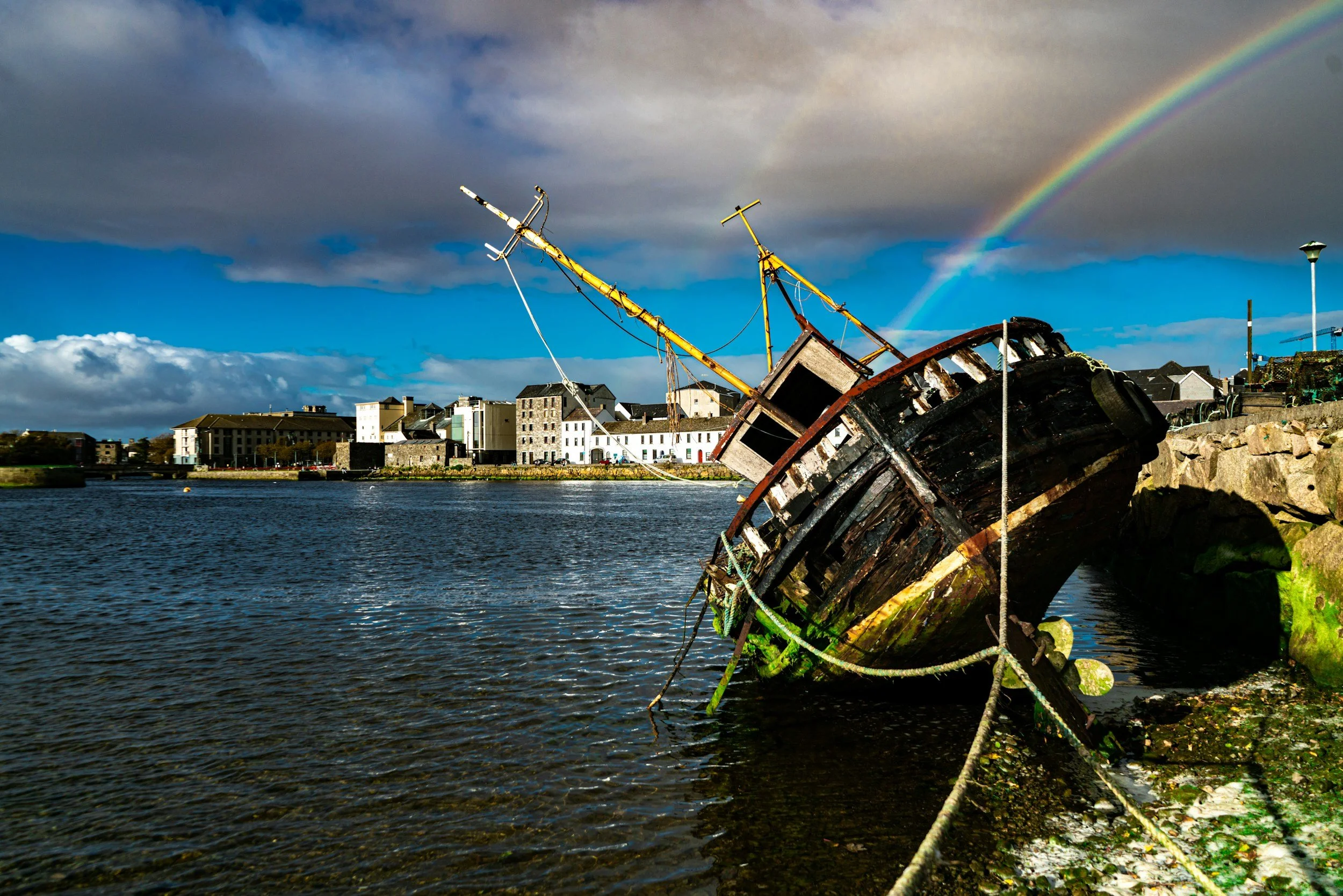 A partially sunken, weathered boat rests against a rocky shoreline with a rainbow in the sky overhead, and modern buildings across the water.