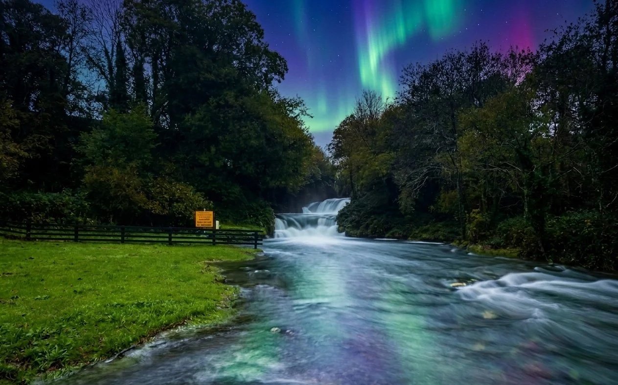 Northern Lights over a river flowing through a forested area at night.