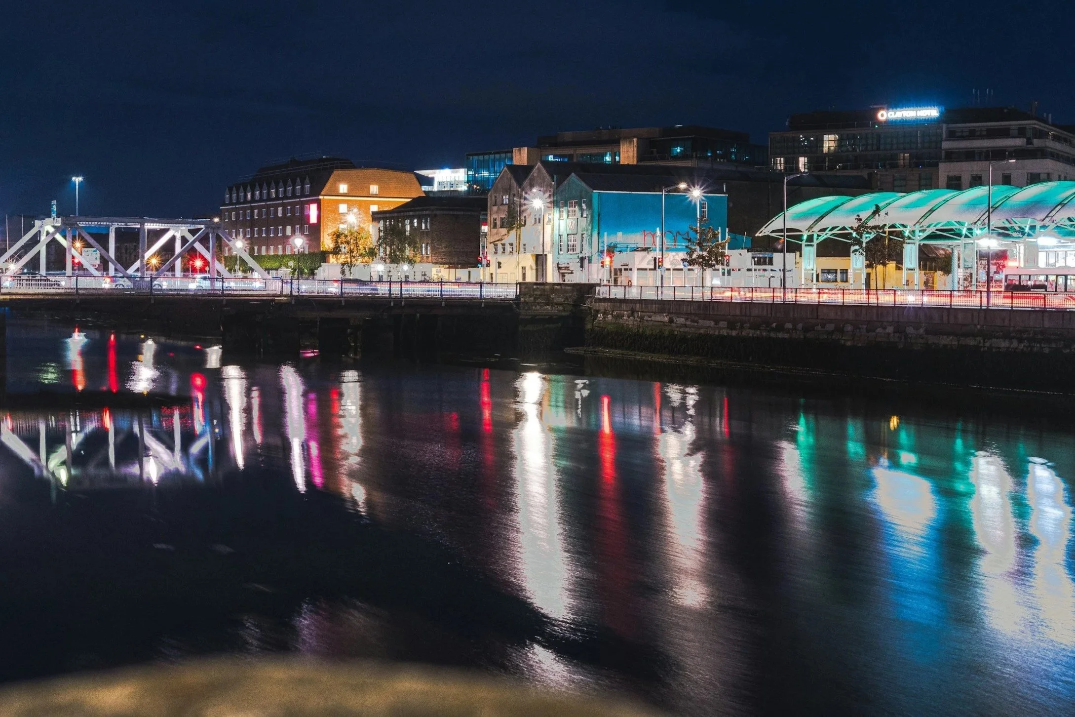 Nighttime cityscape with illuminated buildings and reflections on a river, including a bridge and modern structures with bright lights.