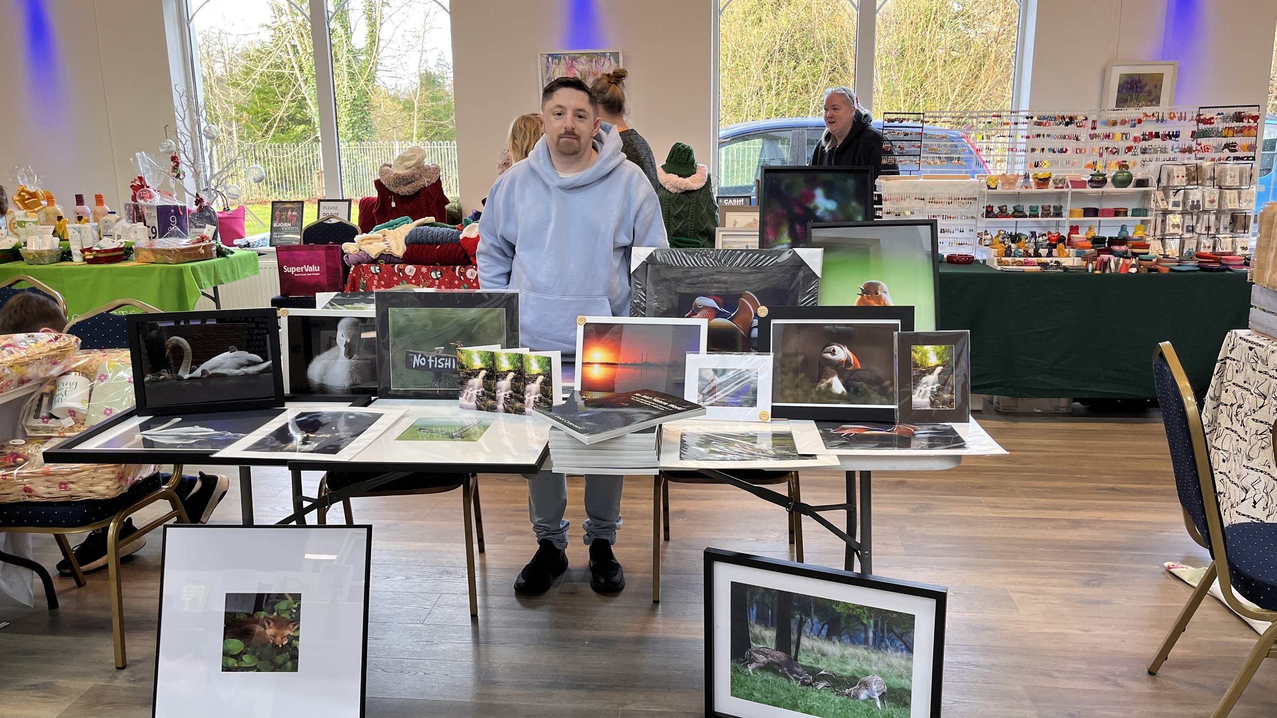 A man stands behind a table displaying framed wildlife photographs, with other tables and booths in the background at an indoor craft fair or market.