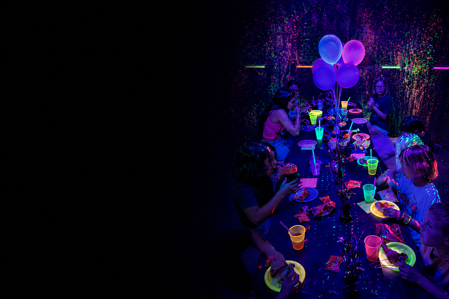 Kids celebrating a birthday party in Pineapple Picasso’s neon Splatter Spot room with glowing plates, cups, balloons, and colorful blacklight paint splatter walls.
