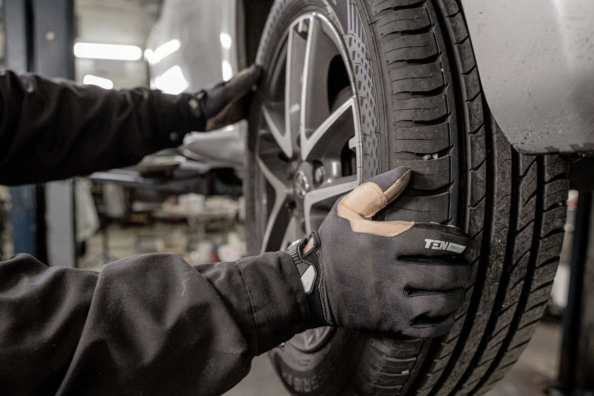 A mechanic wearing black gloves checks a car's tire with a wrench in an auto repair shop.